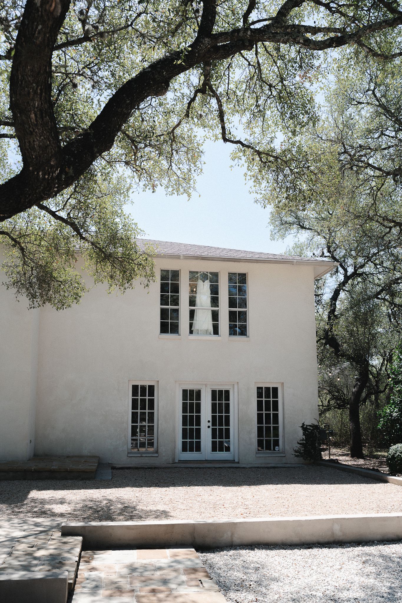 White stucco two-story building with French doors, framed by sprawling oak trees on a gravel courtyard.