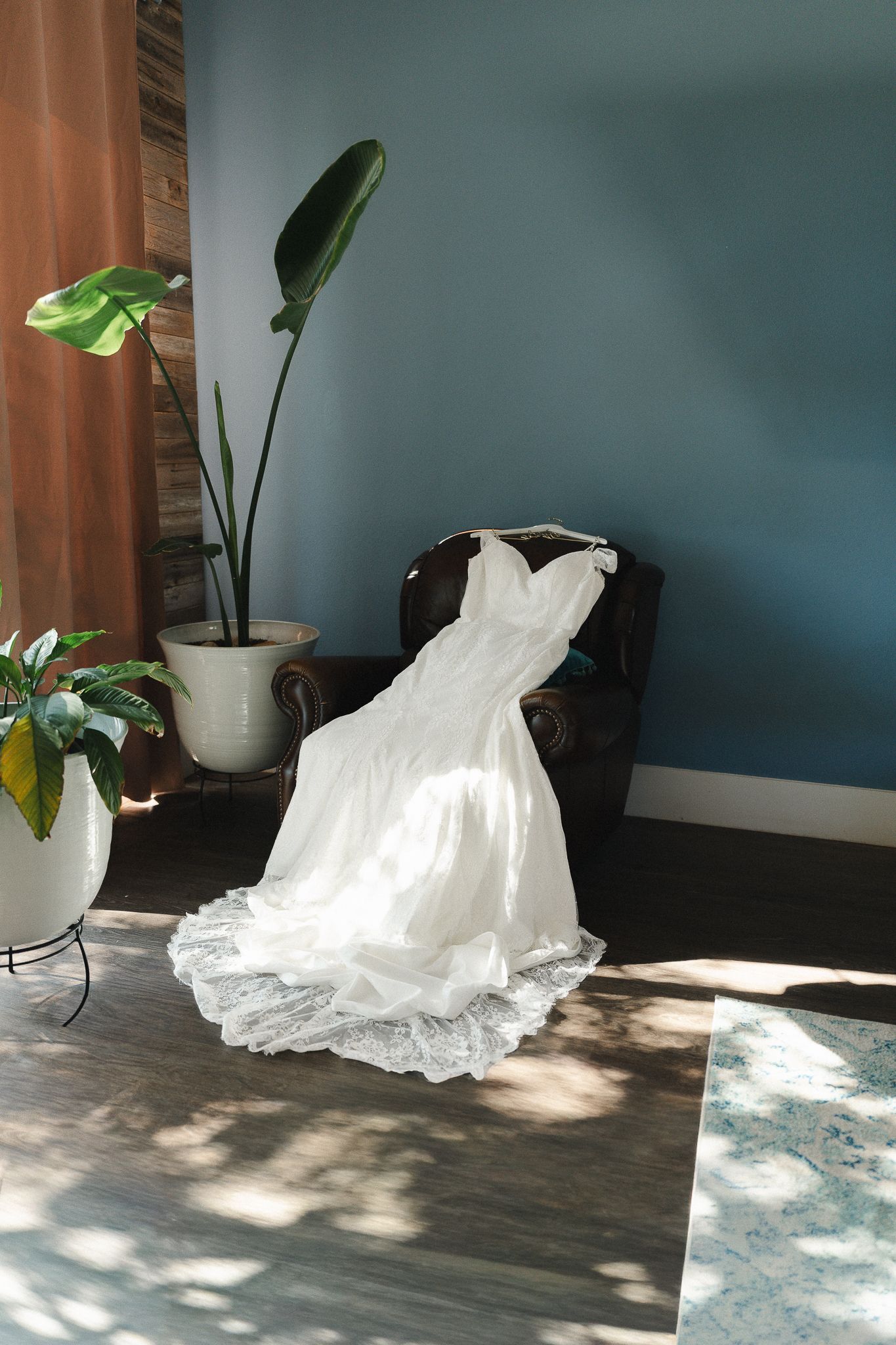 White lace wedding dress draped over a dark chair in a blue room with tropical plants and natural sunlight.