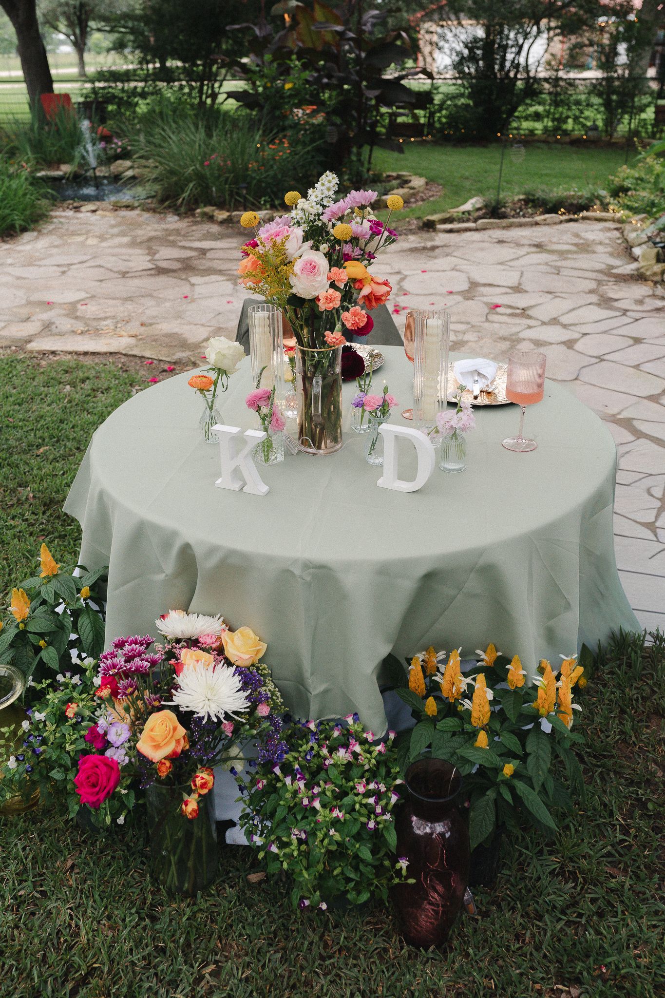 Outdoor wedding sweetheart table with sage green linen, colorful floral arrangements, and monogram letters K and D.