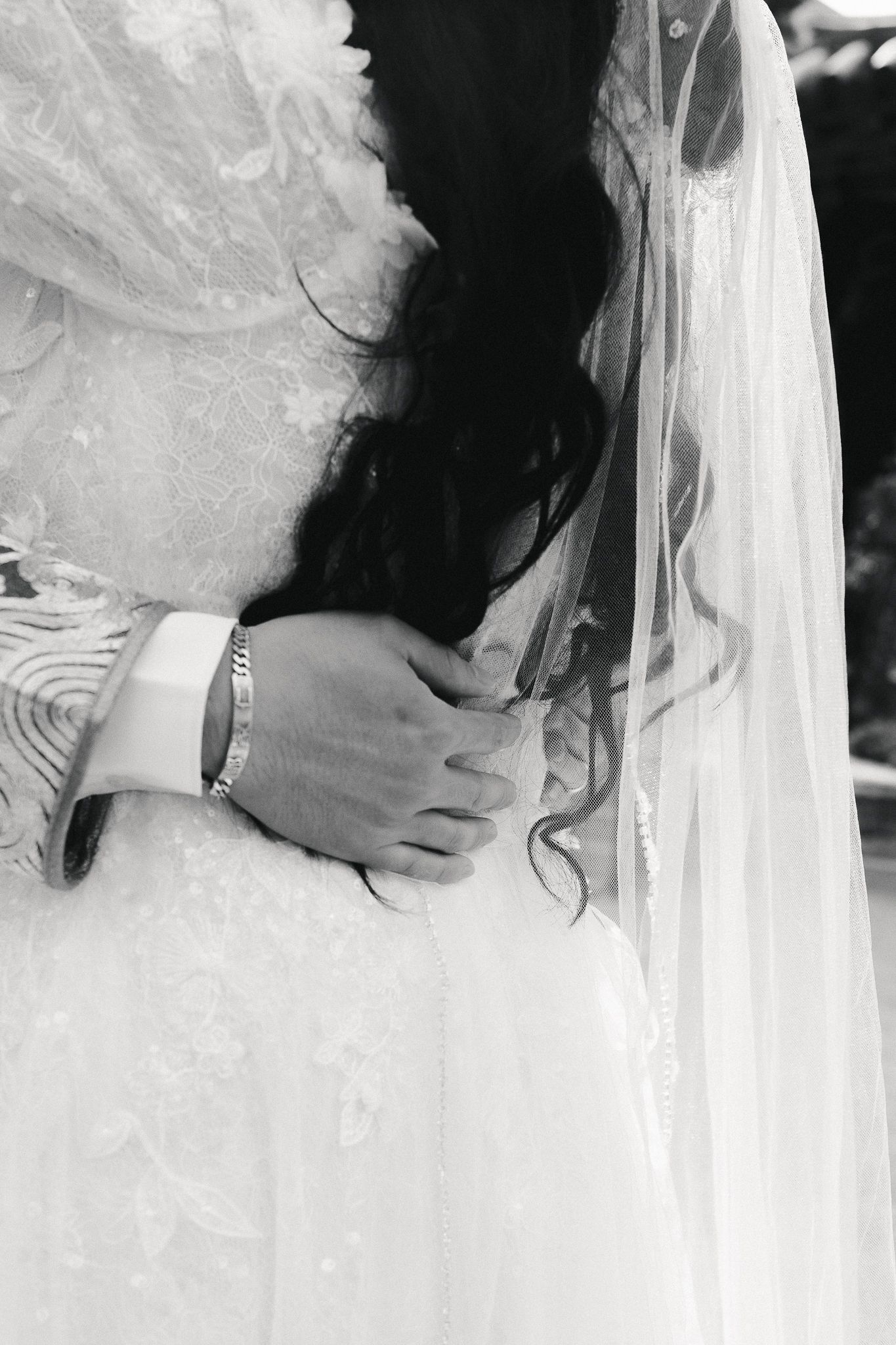 Black and white close-up of groom embracing bride in lace wedding dress with flowing veil, showing bracelet detail.