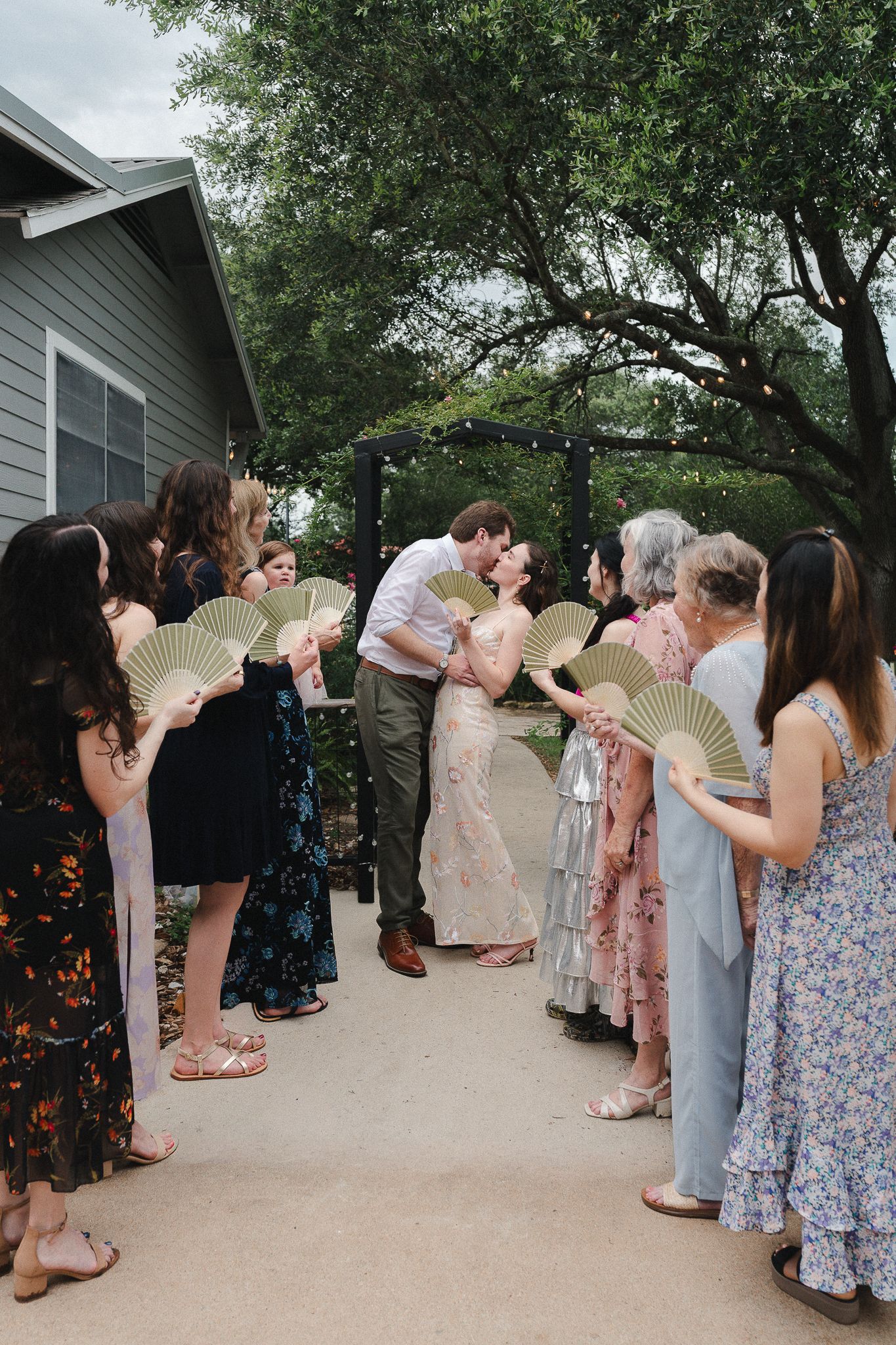Couple shares a kiss while guests hold gold fans lining the aisle at an outdoor wedding ceremony.