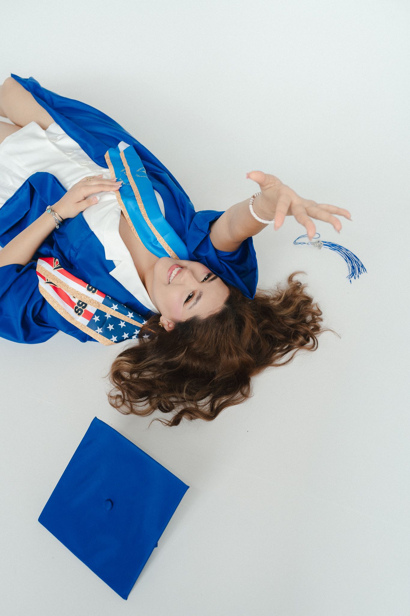 Young woman in blue graduation gown and cap lying on white floor, holding diploma and celebrating academic achievement.