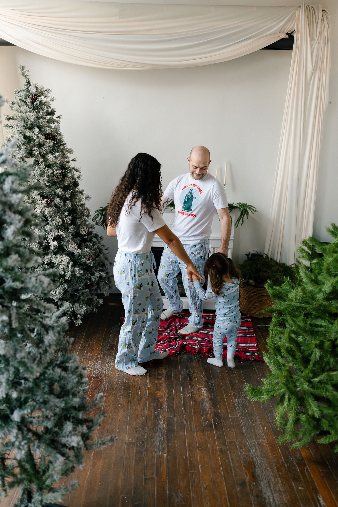 Family in matching Christmas pajamas decorates a frosted artificial tree on rustic wooden floors.