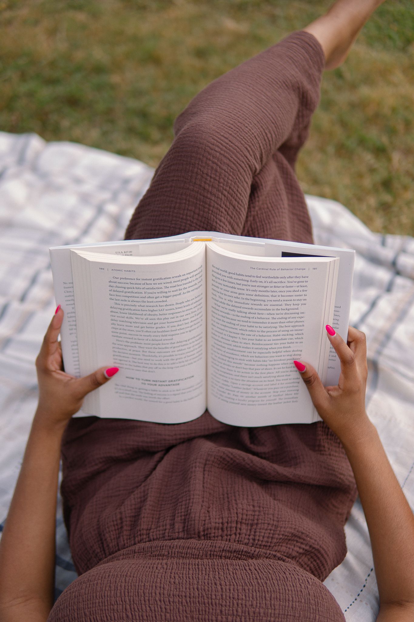 A person relaxes on a blanket while reading a paperback book outdoors on a sunny day.