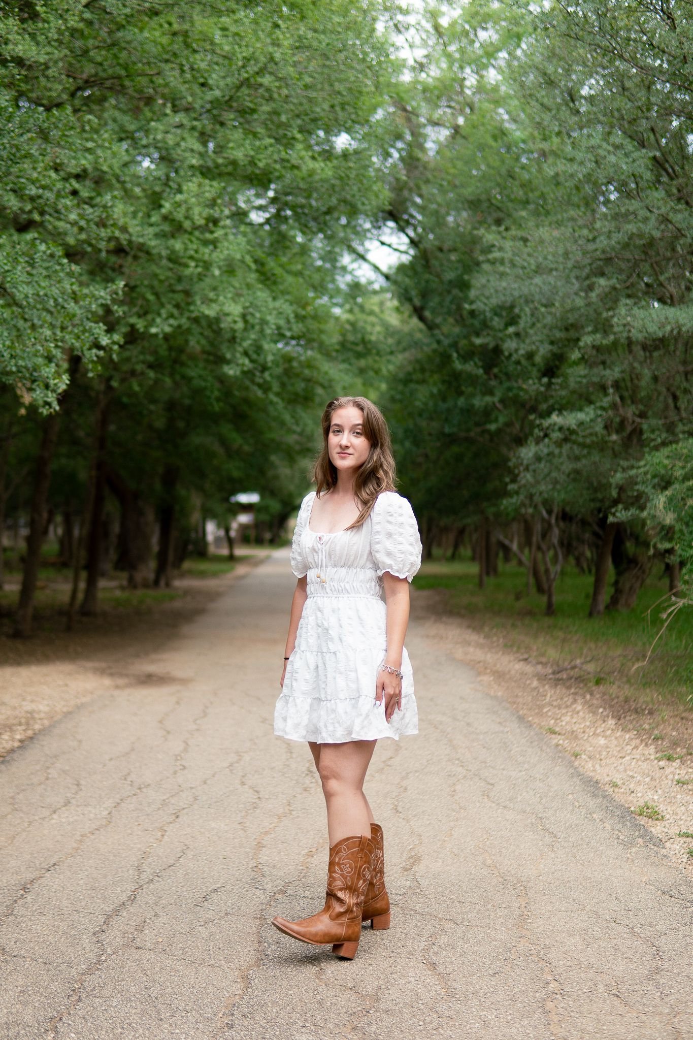 A person wearing a white floral dress and brown cowboy boots poses on a tree-lined dirt path in a rustic outdoor setting.