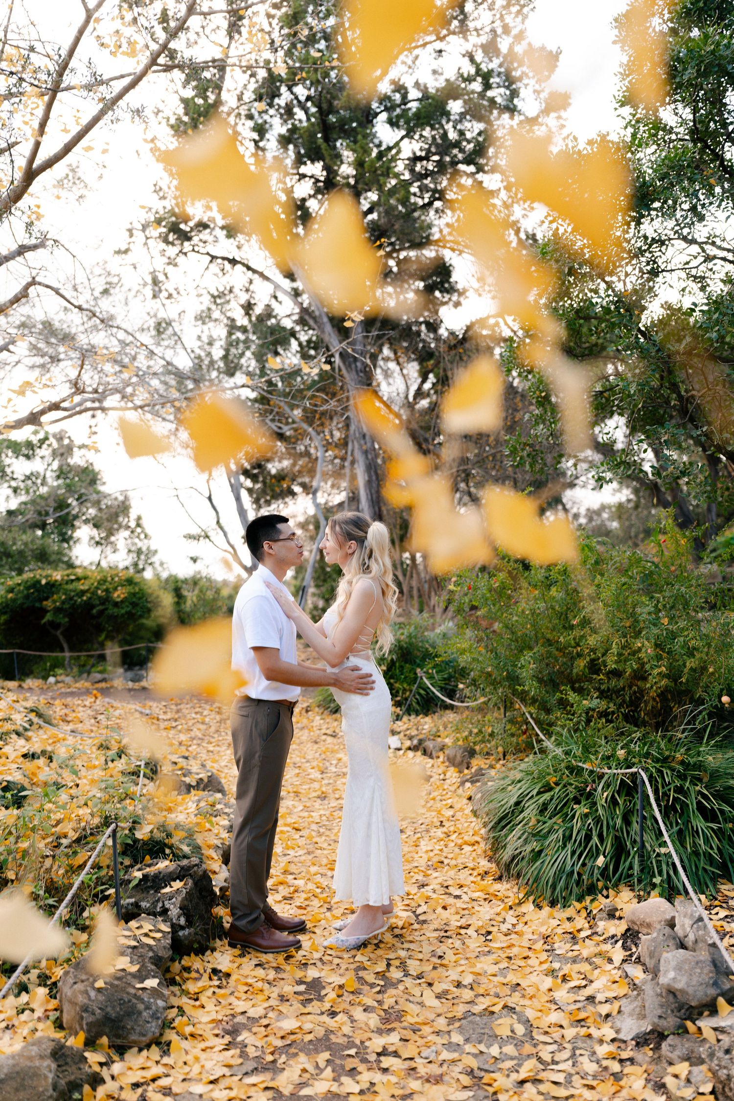 A couple stands together on fallen autumn leaves under trees with golden foliage creating a romantic outdoor scene.