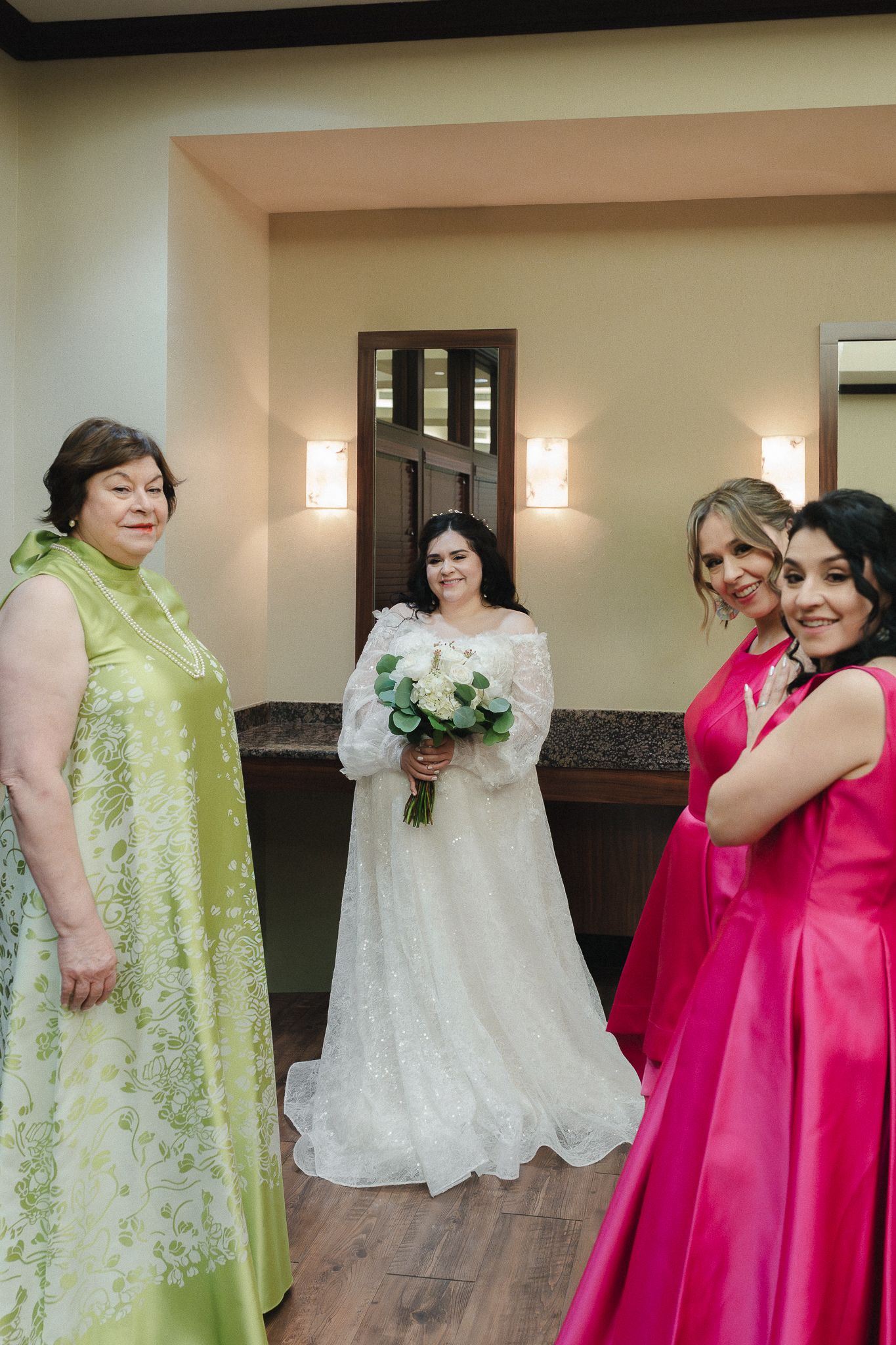 Bride in white gown holding bouquet stands with women in green and pink dresses in elegant wedding venue.