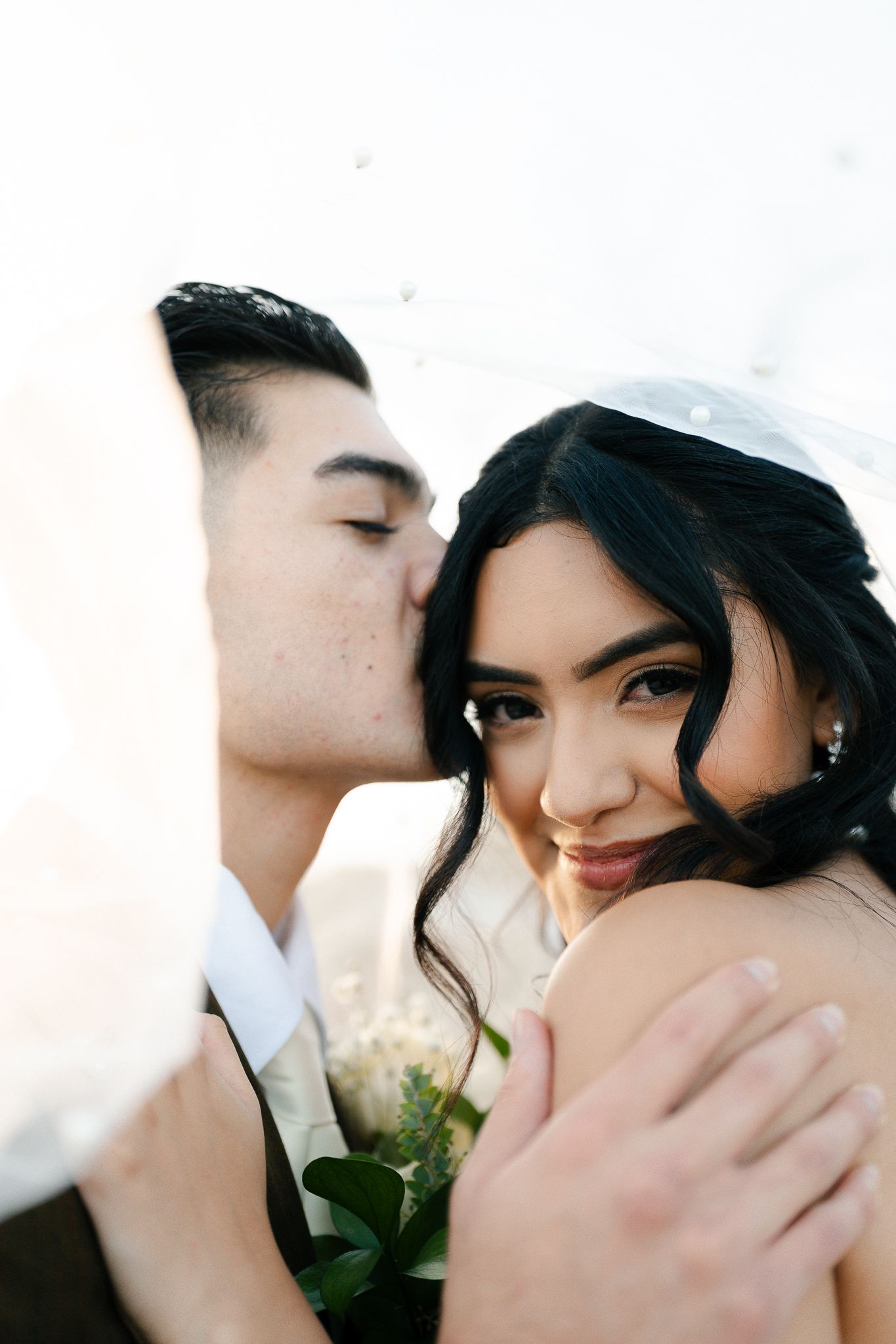 A groom tenderly kisses his bride on the cheek as she smiles, both dressed in wedding attire with a soft bright background.