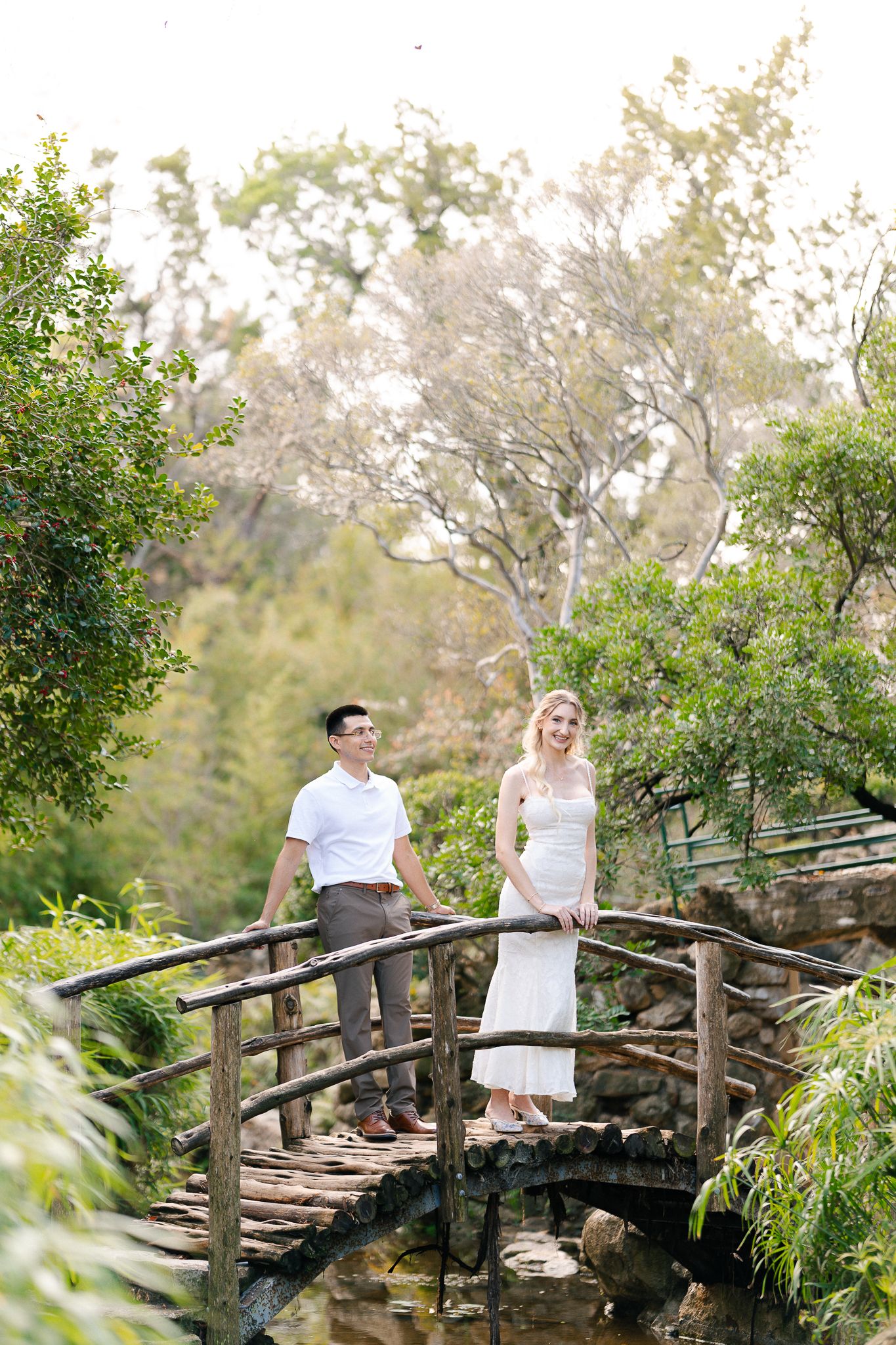 A couple shares an intimate moment on a rustic wooden bridge in a garden setting.