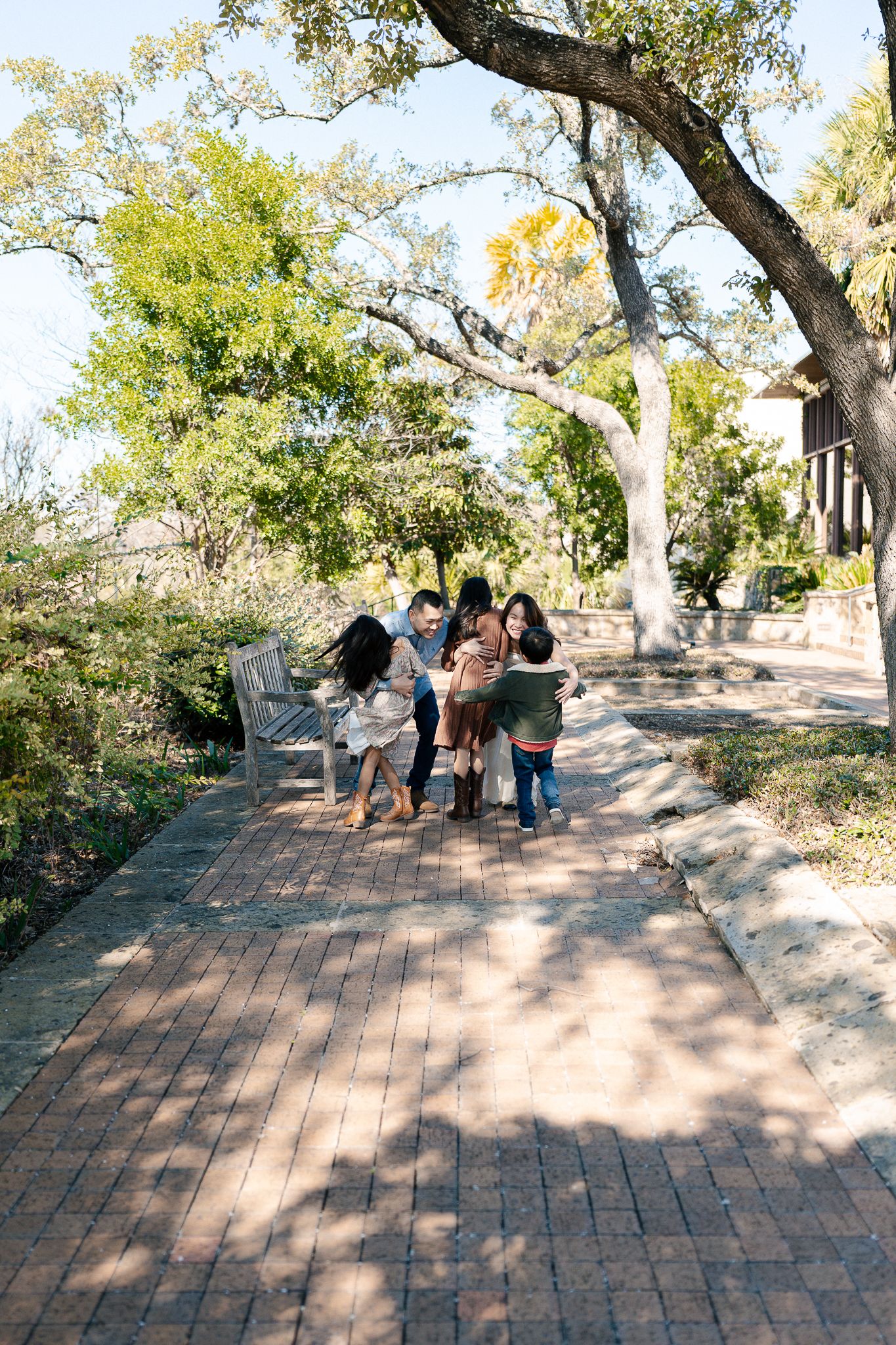 A group of people walking along a curved brick pathway surrounded by trees and dappled sunlight.