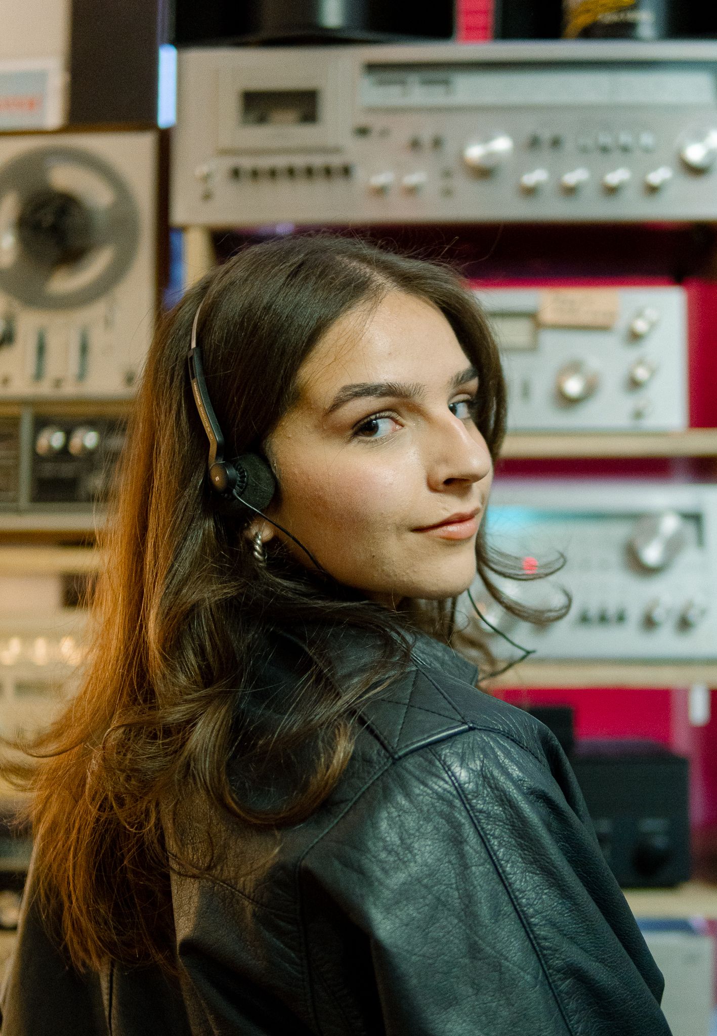 Someone wearing headphones and a leather jacket looks thoughtfully to the side in a room filled with audio equipment.