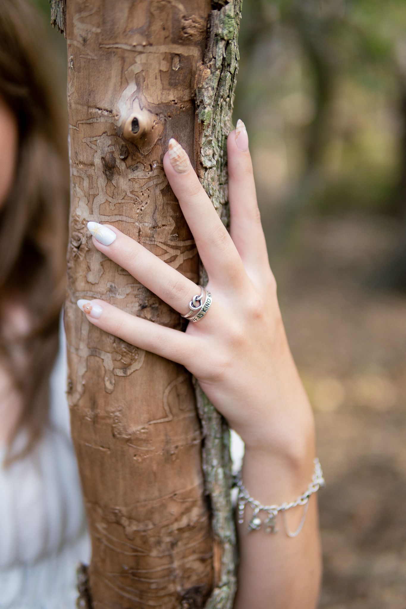 A hand wearing an elegant ring and charm bracelet touches the bark of a tree trunk in a natural outdoor setting.