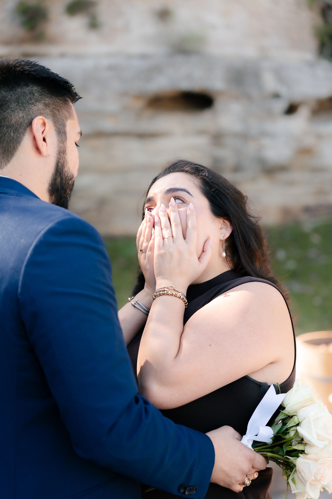 A couple shares an emotional moment during a proposal as she covers her mouth in surprise while he holds white flowers.