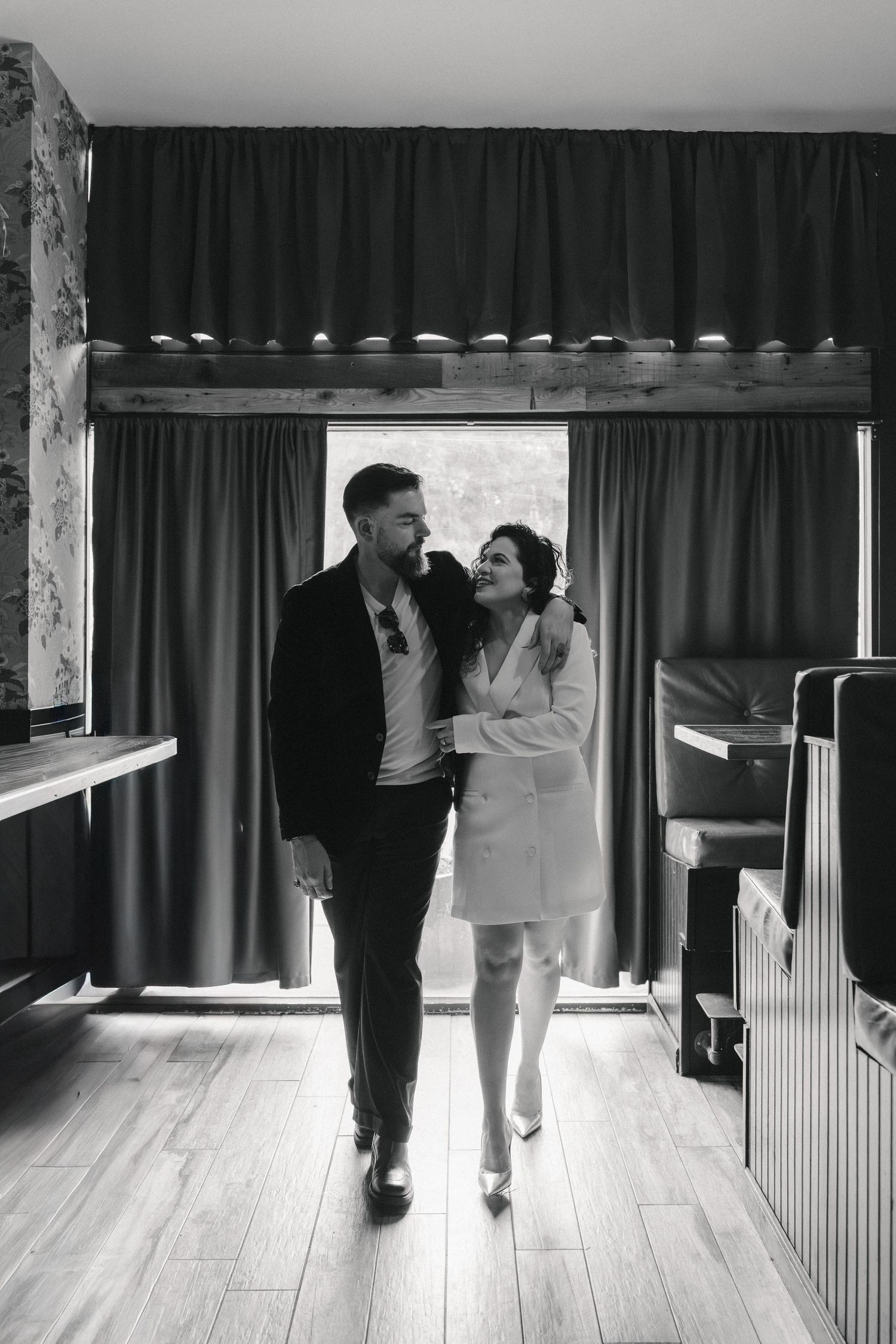 Black and white photo of an elegant couple walking arm in arm through a stylish room with curtains and wooden floors.