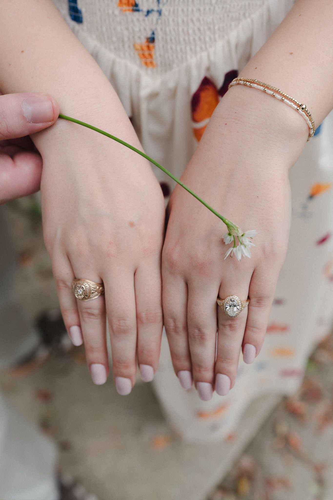 Two hands adorned with gold rings and a bracelet holding a delicate white wildflower stem against a floral dress.