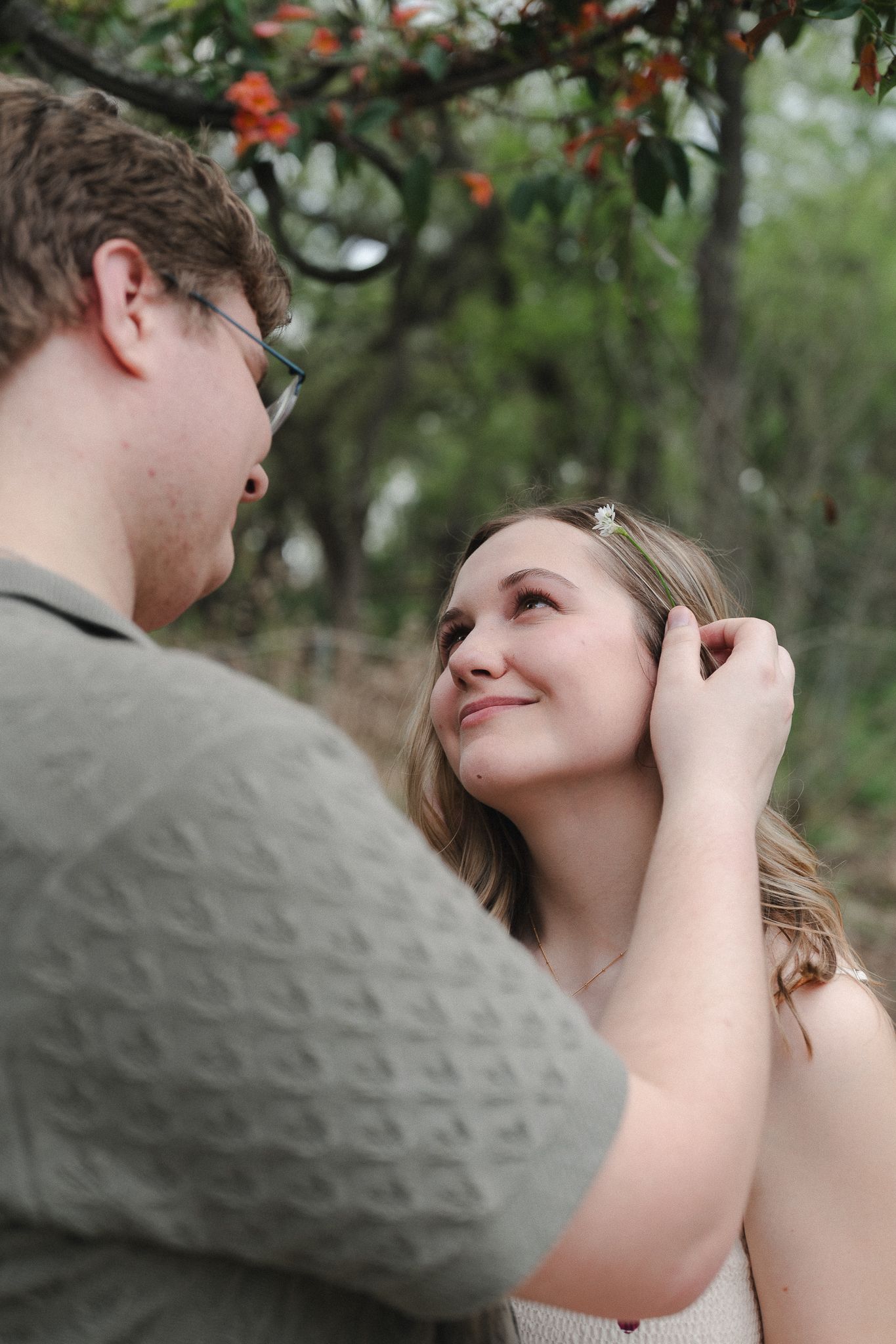A young couple shares a tender moment outdoors, as a man gently touches a smiling woman's hair beneath flowering trees.