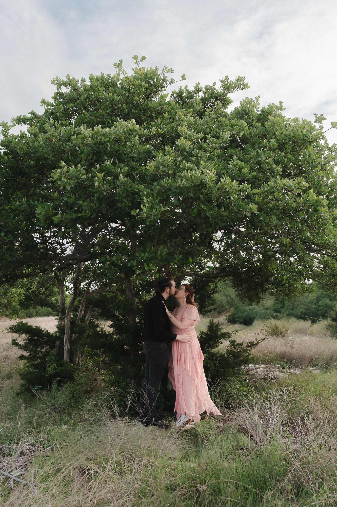 Couple kissing under a lush green tree in a field, woman wearing a flowing pink dress in a romantic outdoor setting.