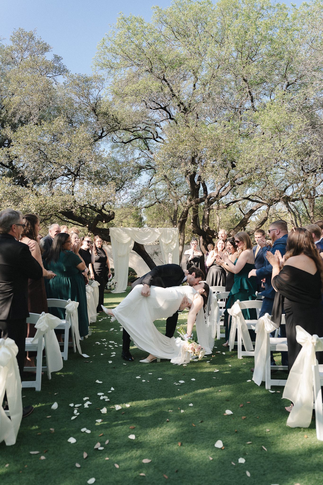 Bride and groom share a romantic dip kiss at outdoor wedding ceremony with white chairs and rose petals on green lawn.