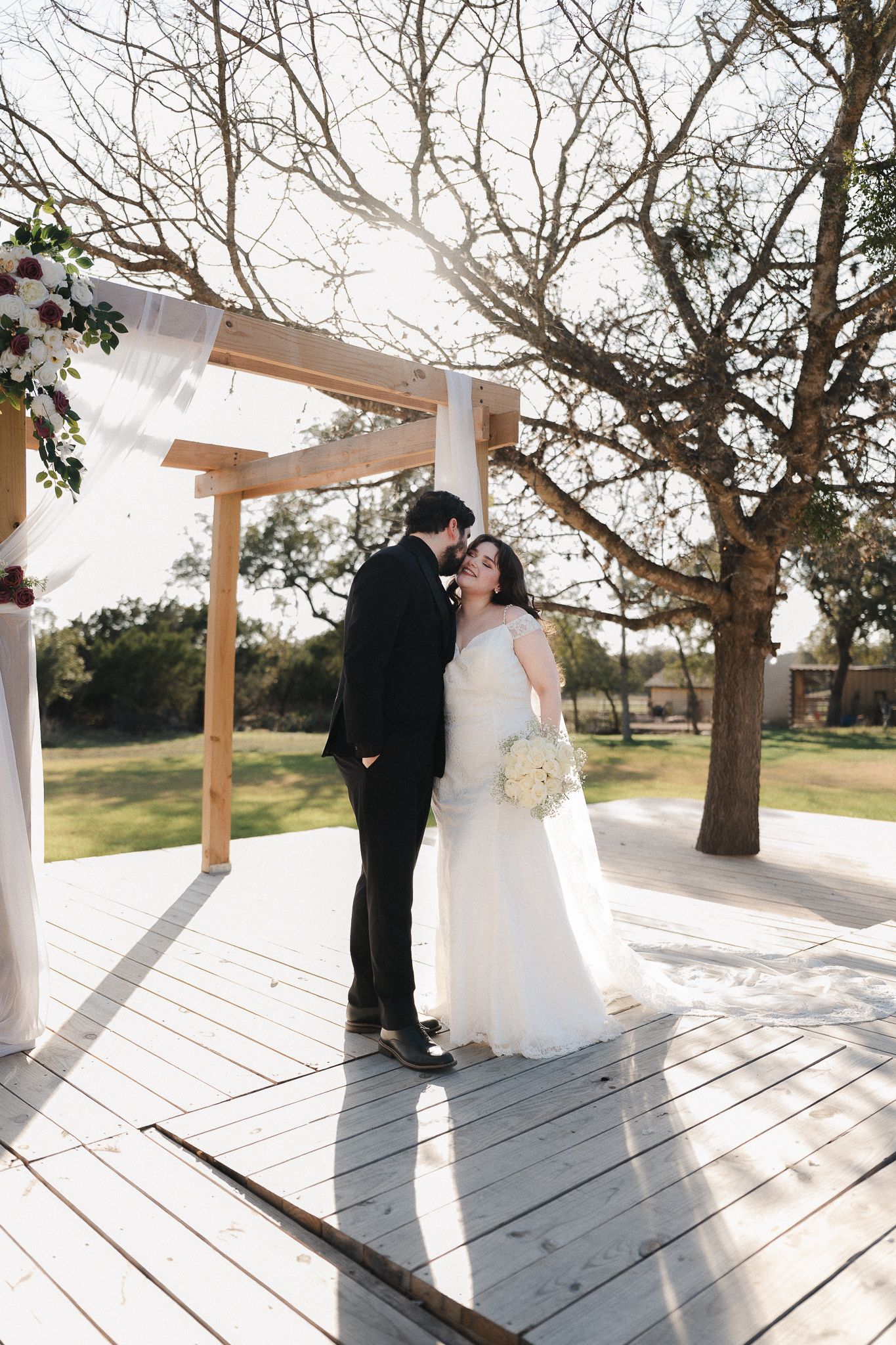 Bride and groom share a romantic kiss under a wooden arbor on an outdoor deck surrounded by trees.