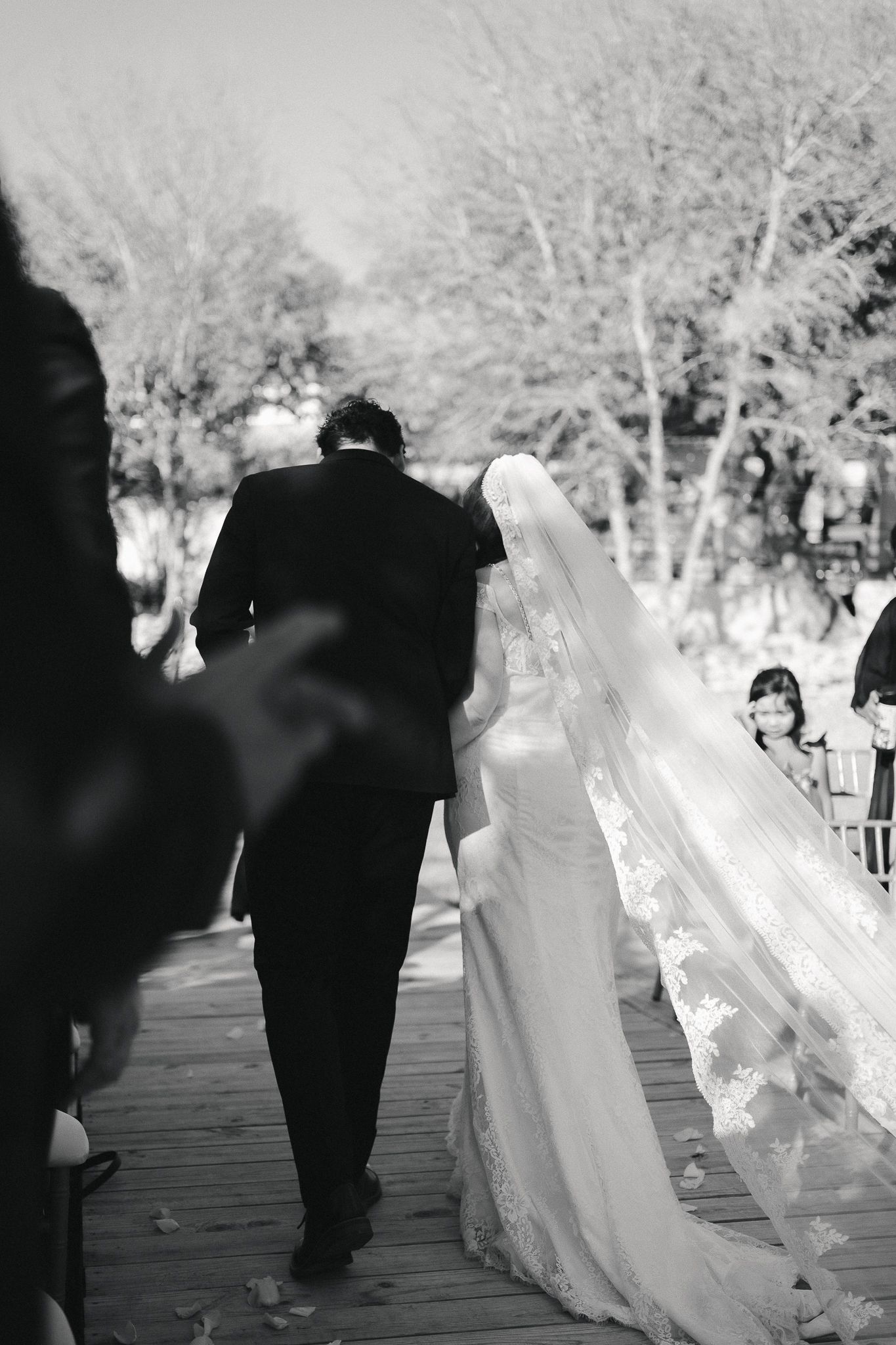 Black and white photo of bride in lace veil walking with groom on wooden path at outdoor wedding ceremony.