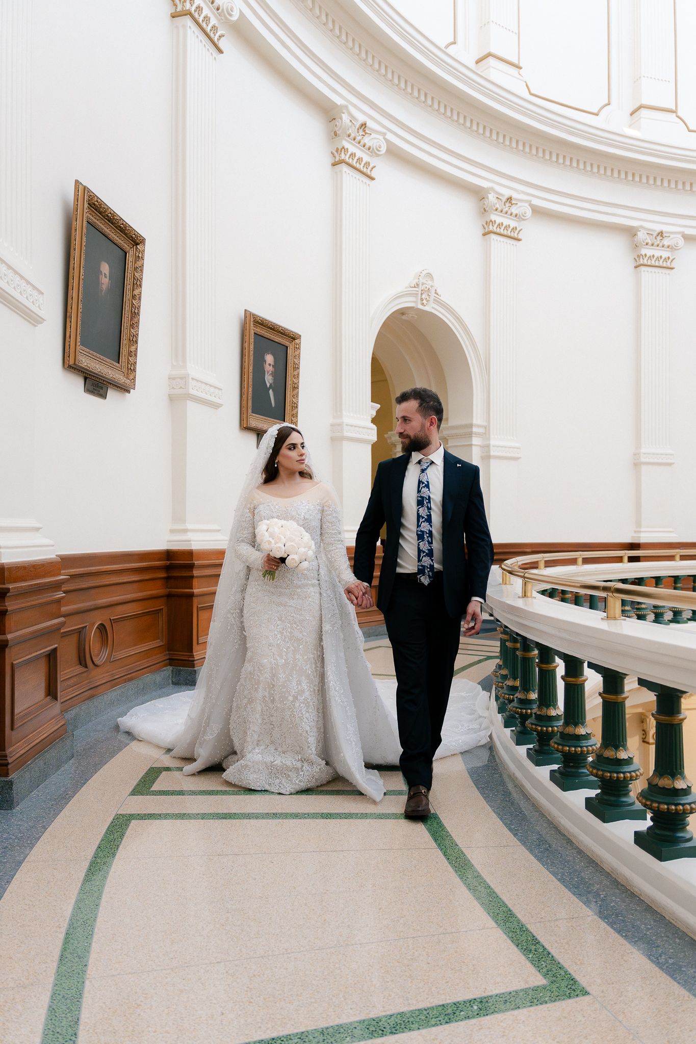 A bride and groom stand in an elegant white curved hallway with ornate architectural details and green-trimmed carpet.
