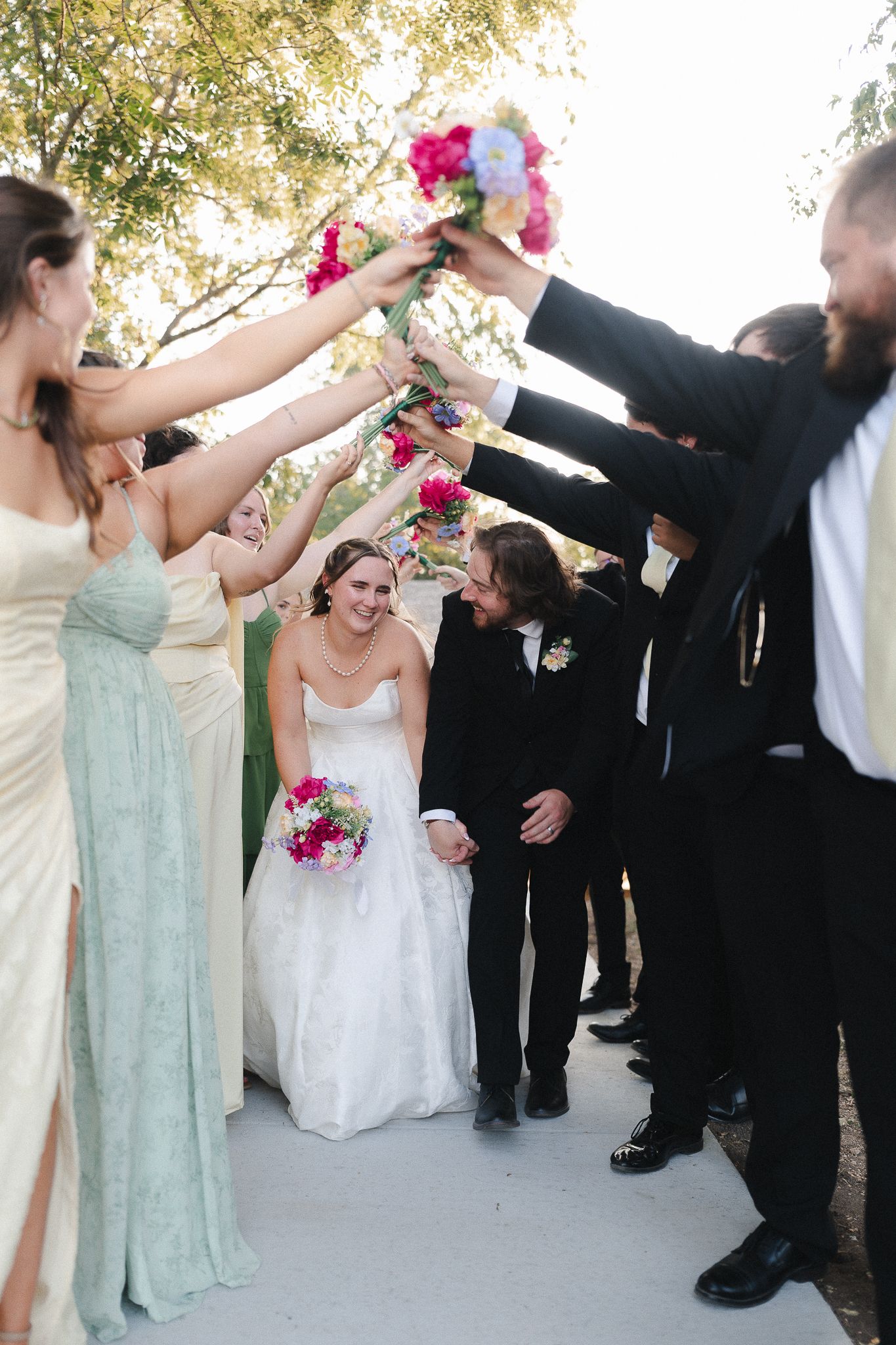 Wedding party raises bouquets in arch as smiling bride and groom walk through tunnel of flowers outdoors.