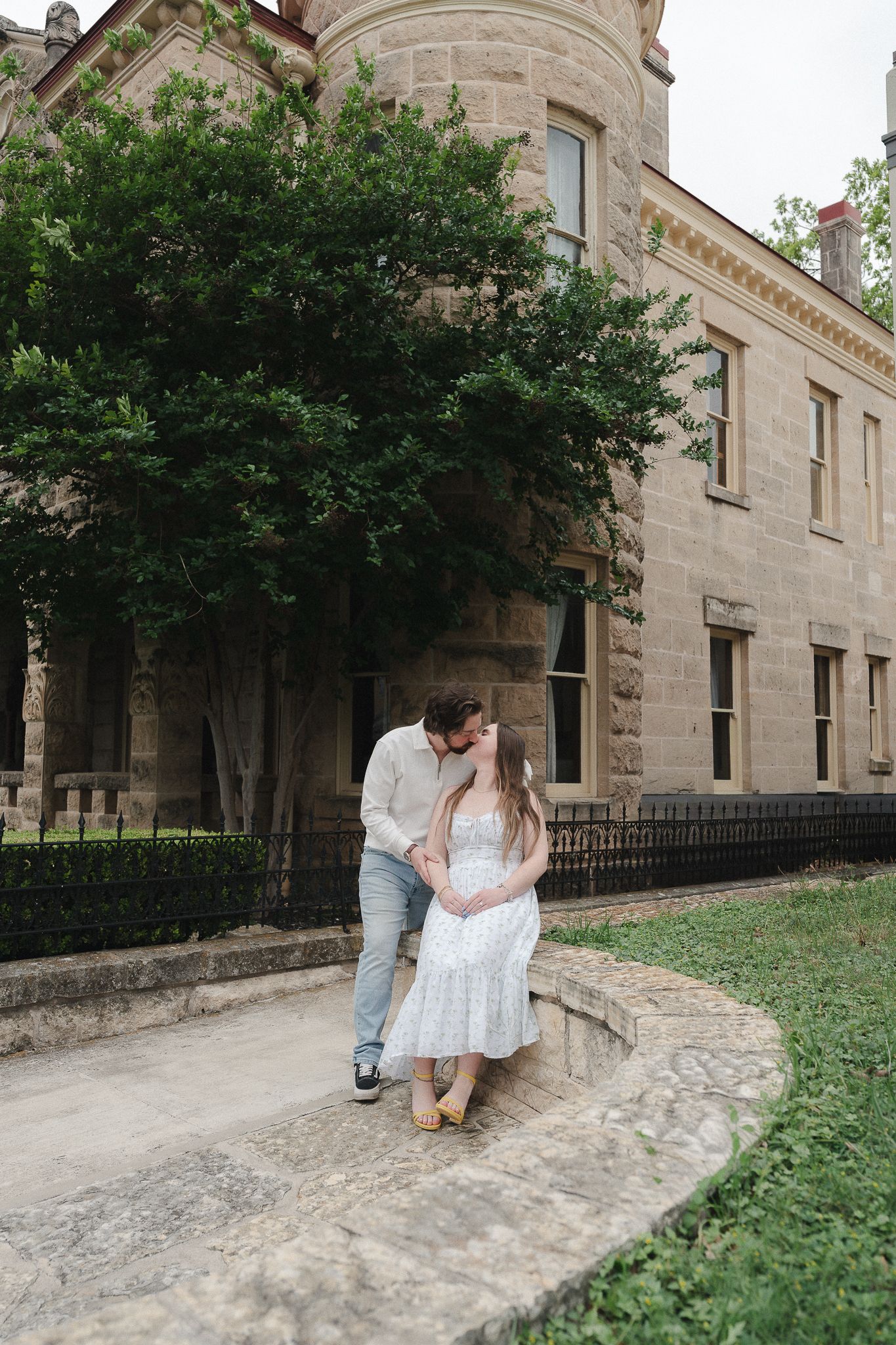 Couple sharing a kiss outside a historic stone building surrounded by lush green trees on a curved pathway.