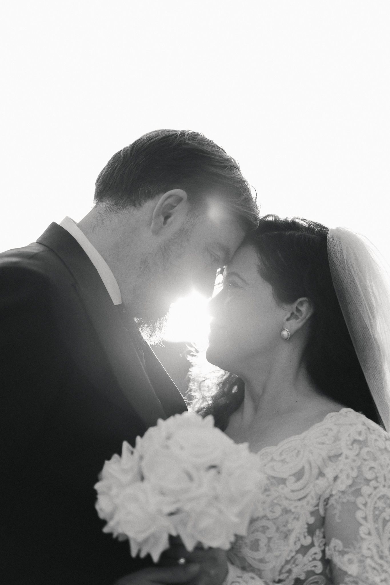 Black and white wedding portrait of a bride and groom touching foreheads, backlit by bright sunlight, with the bride holding a rose bouquet.