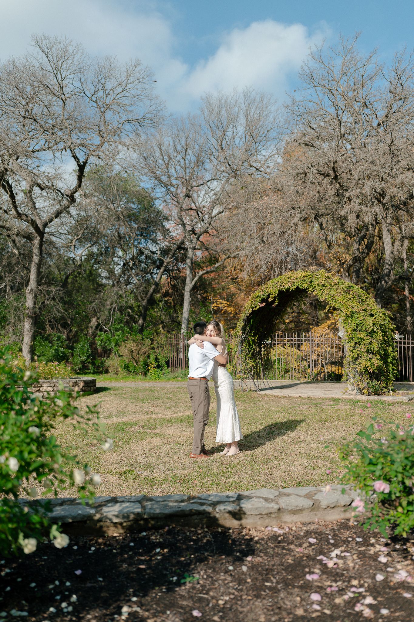 A couple shares a romantic moment in a garden setting with a moss-covered archway and bare winter trees in the background.