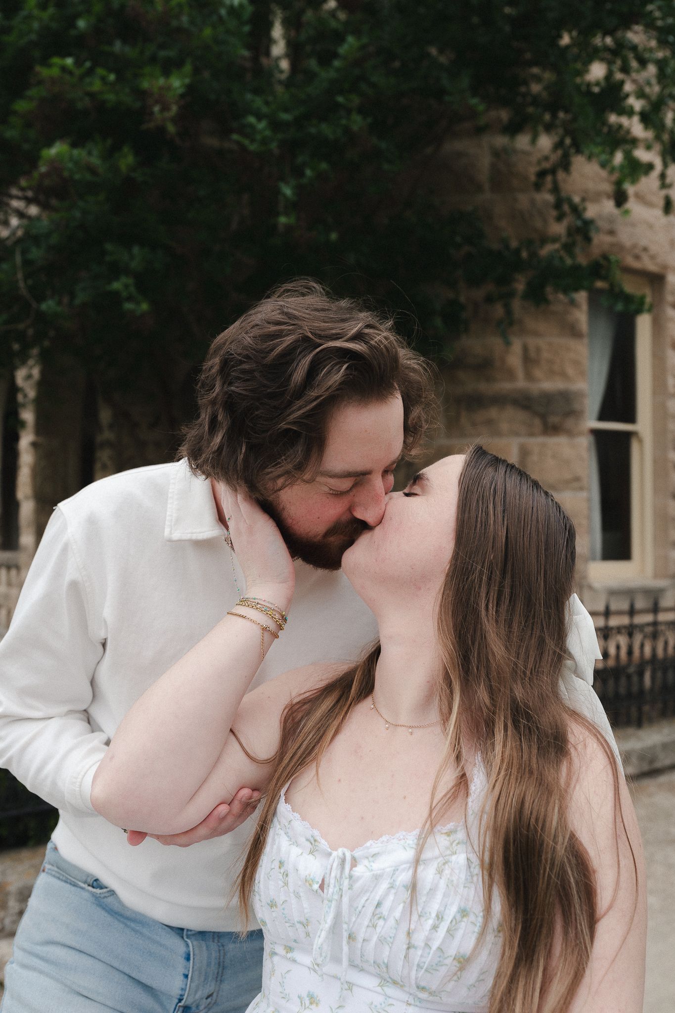 A couple shares a romantic kiss outdoors, with the woman tenderly holding the man's face near a stone building.