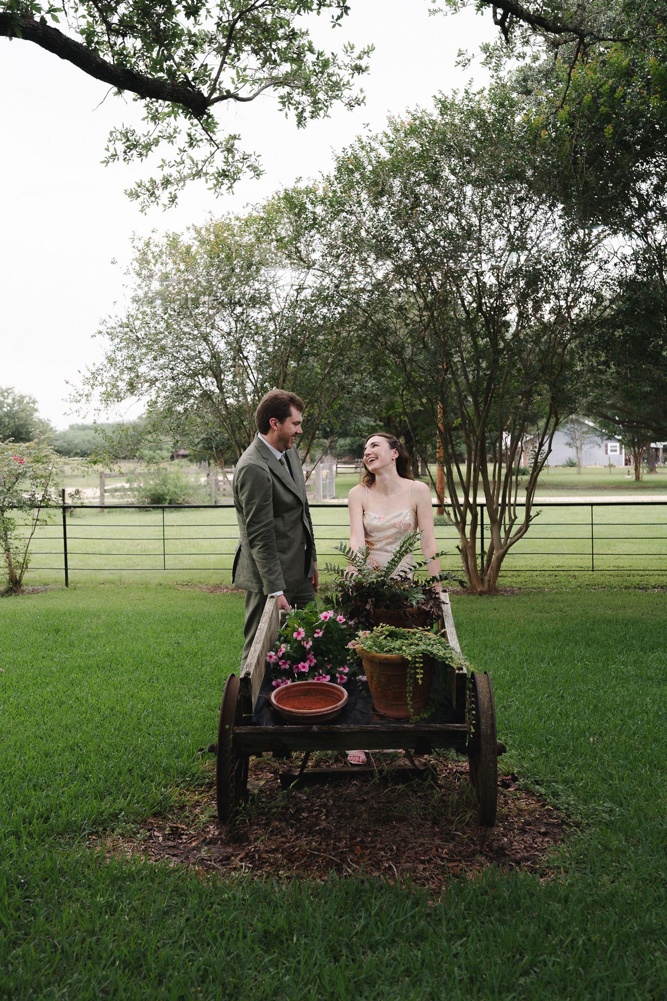A couple smiles at each other beside a rustic wooden cart with potted plants on a lush green lawn.