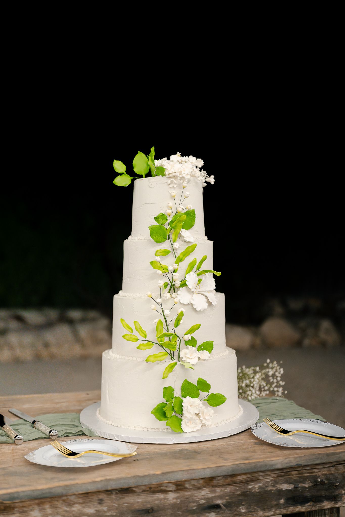Elegant four-tier white wedding cake decorated with fresh green leaves and delicate white flowers on rustic wooden table.