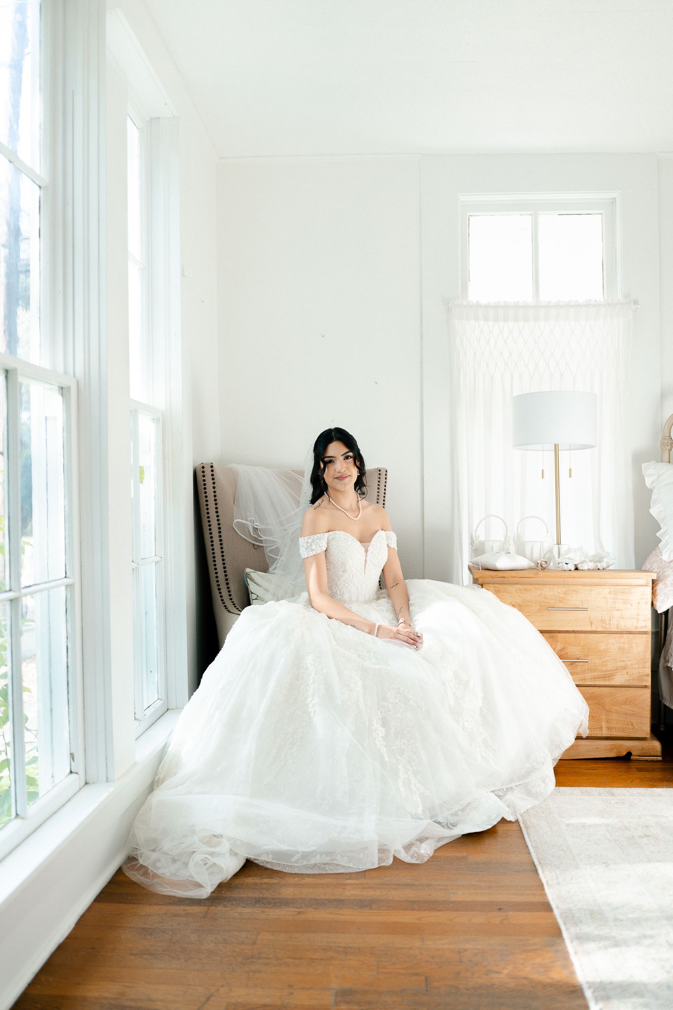 A bride in an elegant off-shoulder white ball gown sits gracefully by a bright window in a minimalist room with hardwood floors.