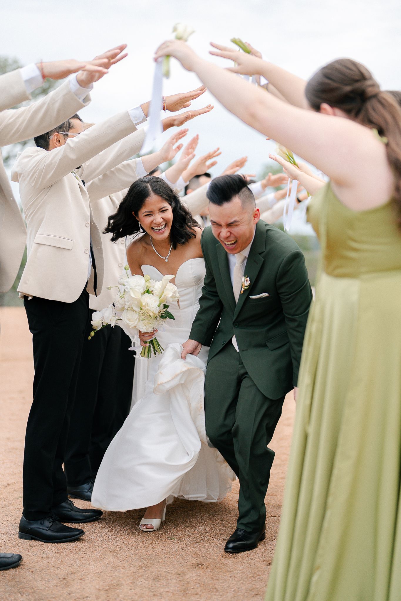 Wedding party celebrates with raised hands as newlyweds walk under their arch.