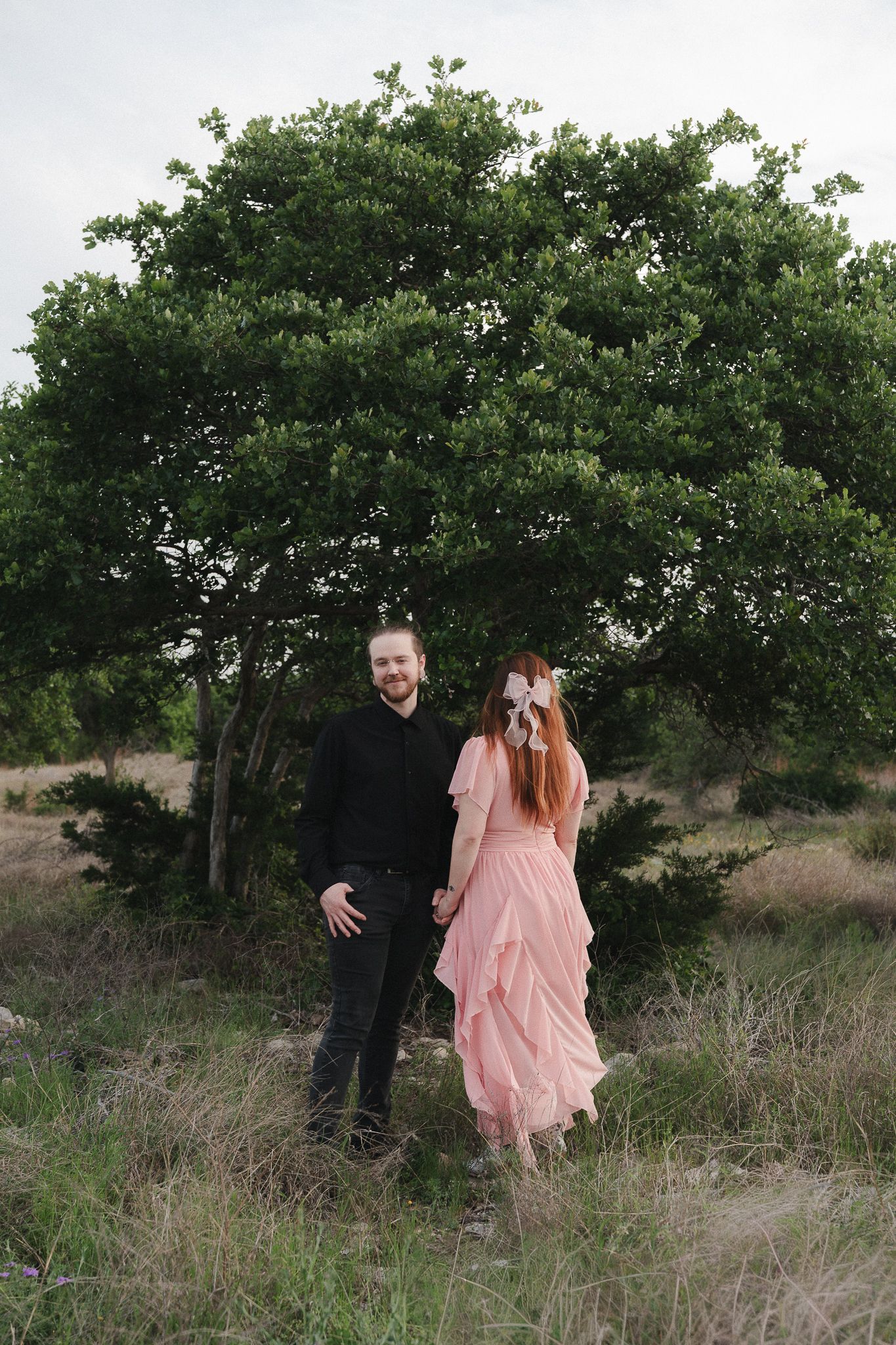 Couple holding hands in a field, man in black outfit and woman in pink ruffled dress, standing before a lush green tree.