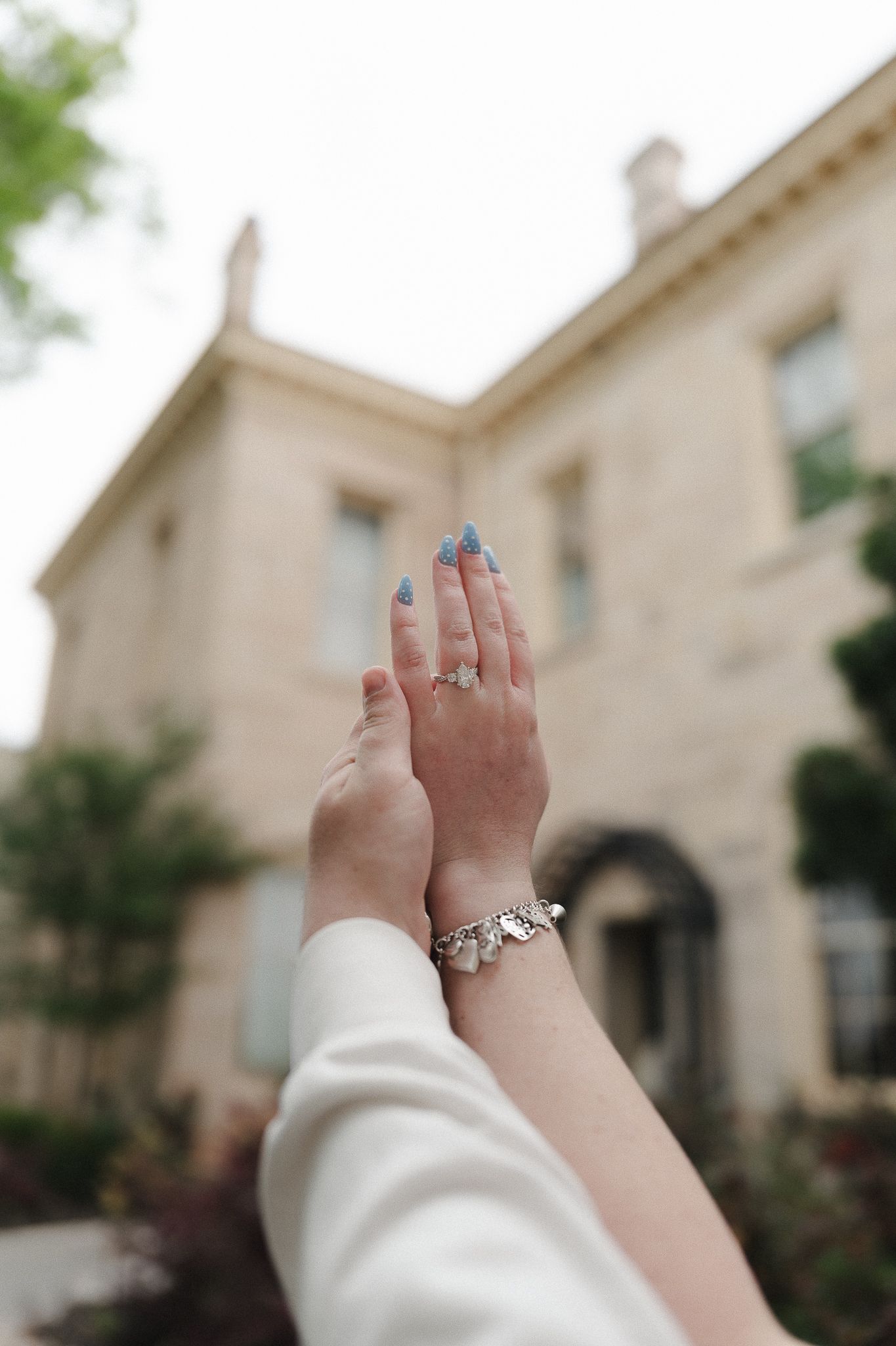 Close-up of hands with blue nails, diamond ring, and silver charm bracelet raised in front of a stone mansion.