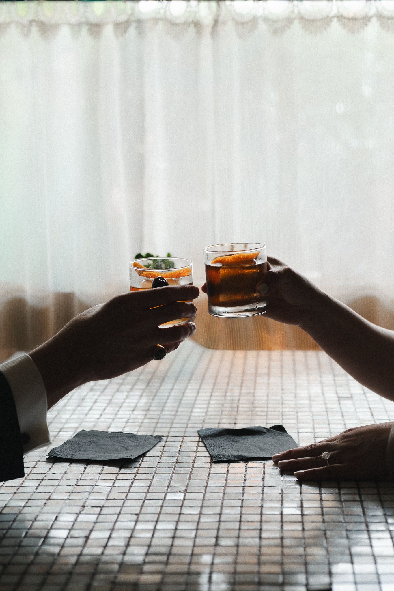Couple toasting cocktails over mosaic tile table, one drink with fruit garnish and one amber whiskey.