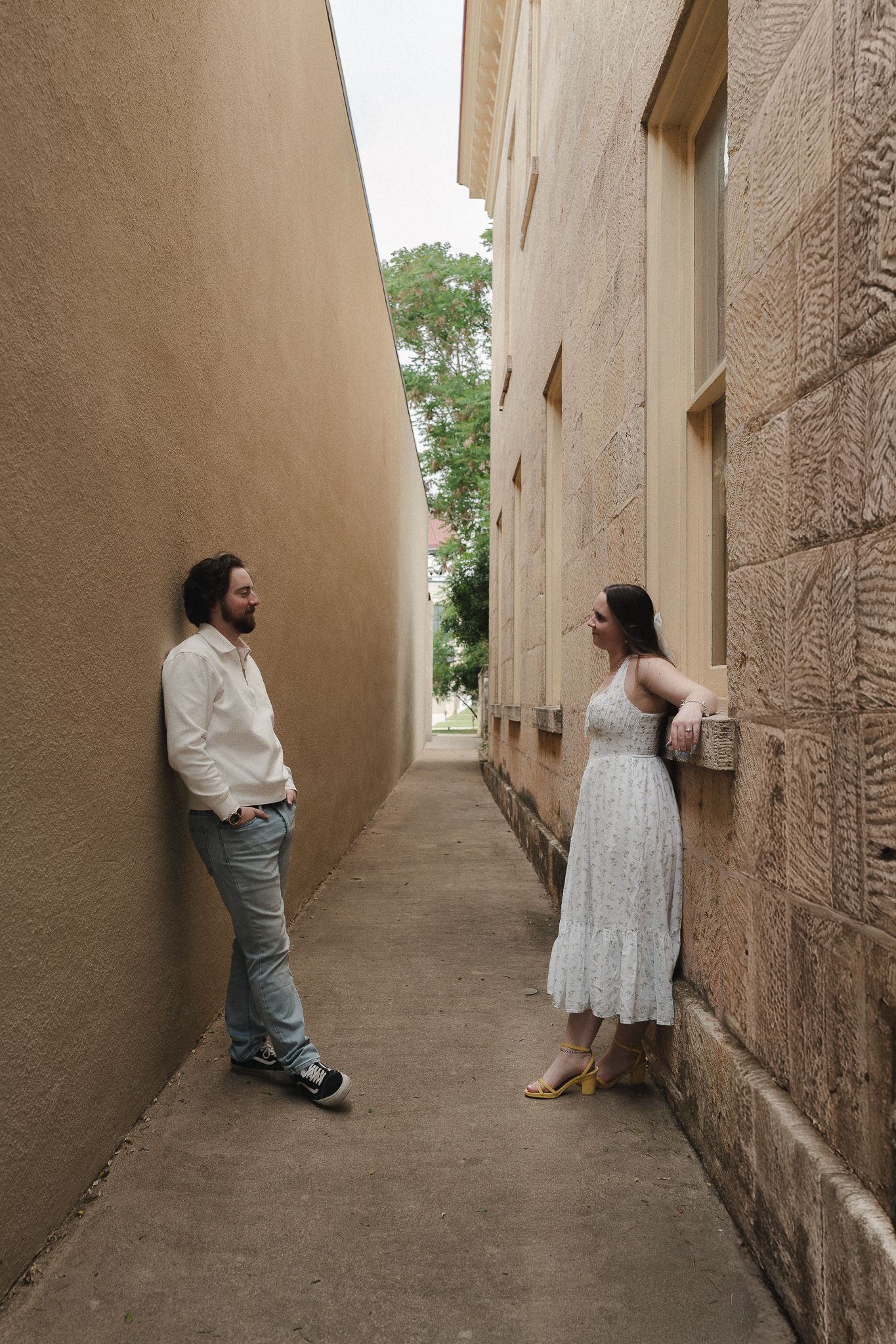 A man and woman stand facing each other in a narrow alley between tan walls, she wears a white dress, he wears jeans.