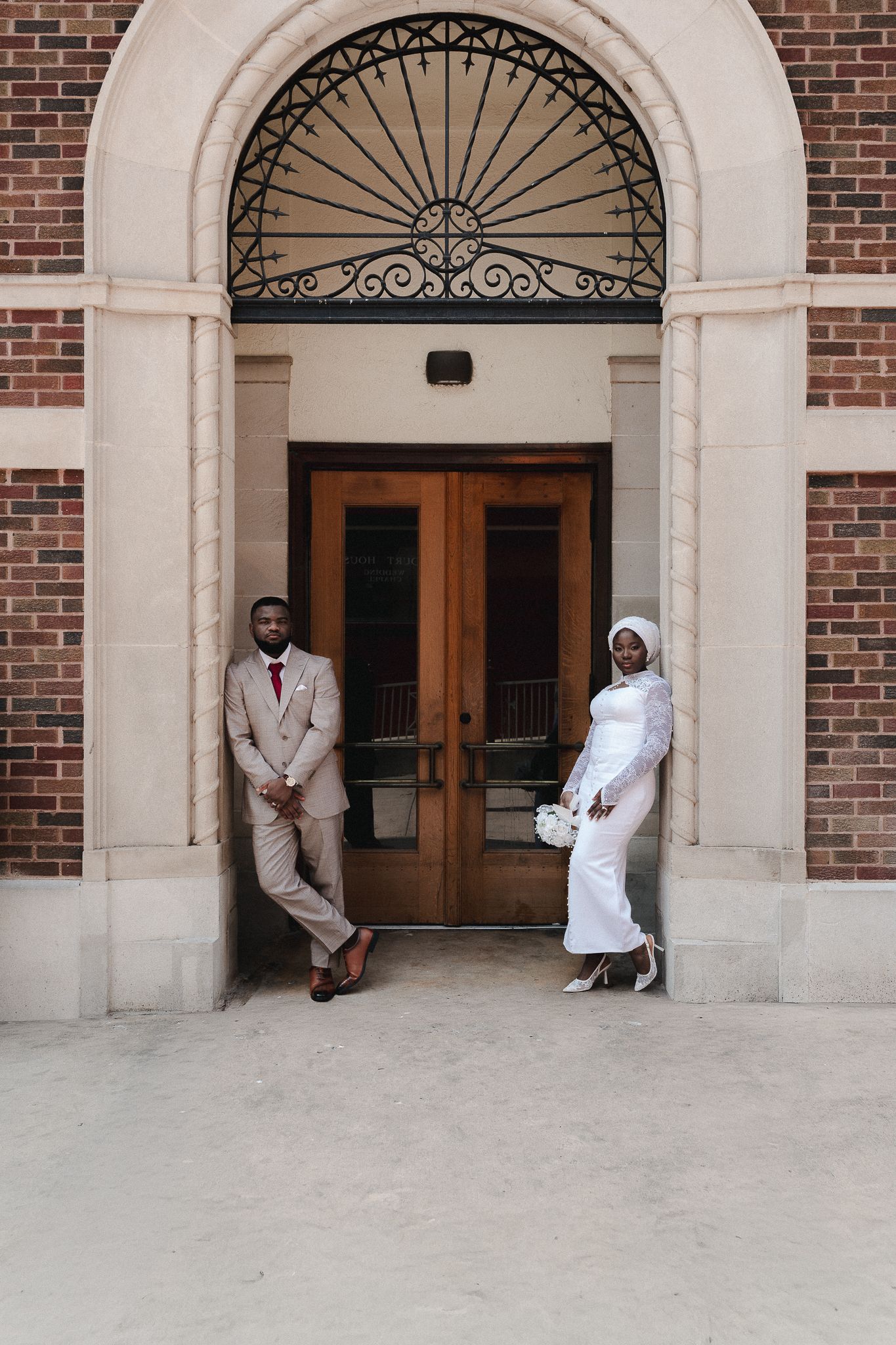 Bride in white hijab gown and groom in tan suit pose at ornate brick building entrance with wooden doors.
