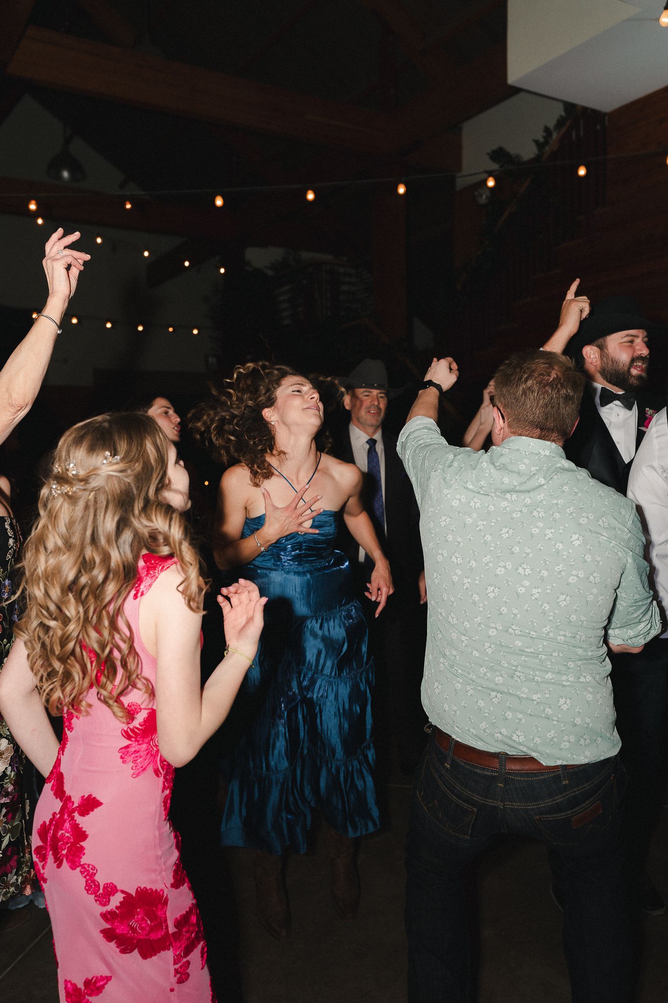 Guests dancing joyfully at a wedding reception under string lights, wearing formal attire including blue dress and floral pink gown.
