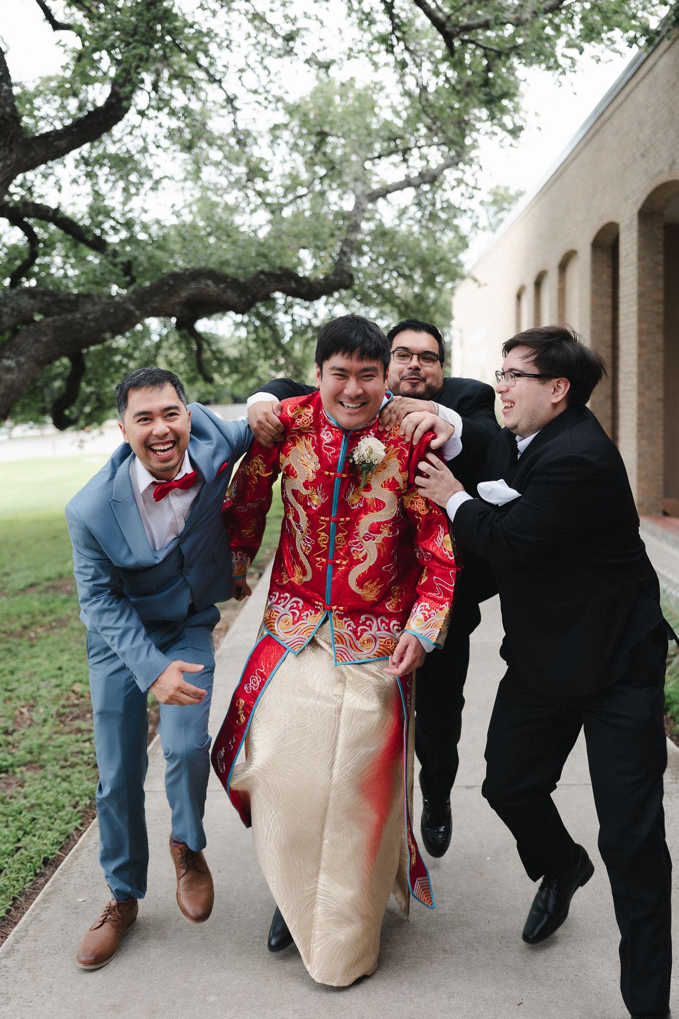 Groom in traditional red Chinese wedding attire laughs as groomsmen playfully lift him on a sidewalk outdoors.