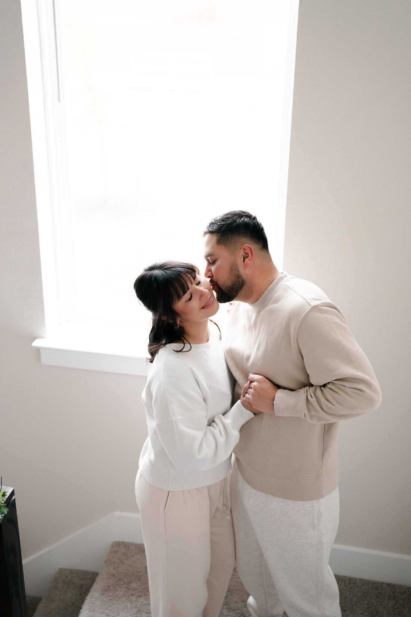 A couple wearing neutral beige and white outfits embrace tenderly in a bright minimalist room.