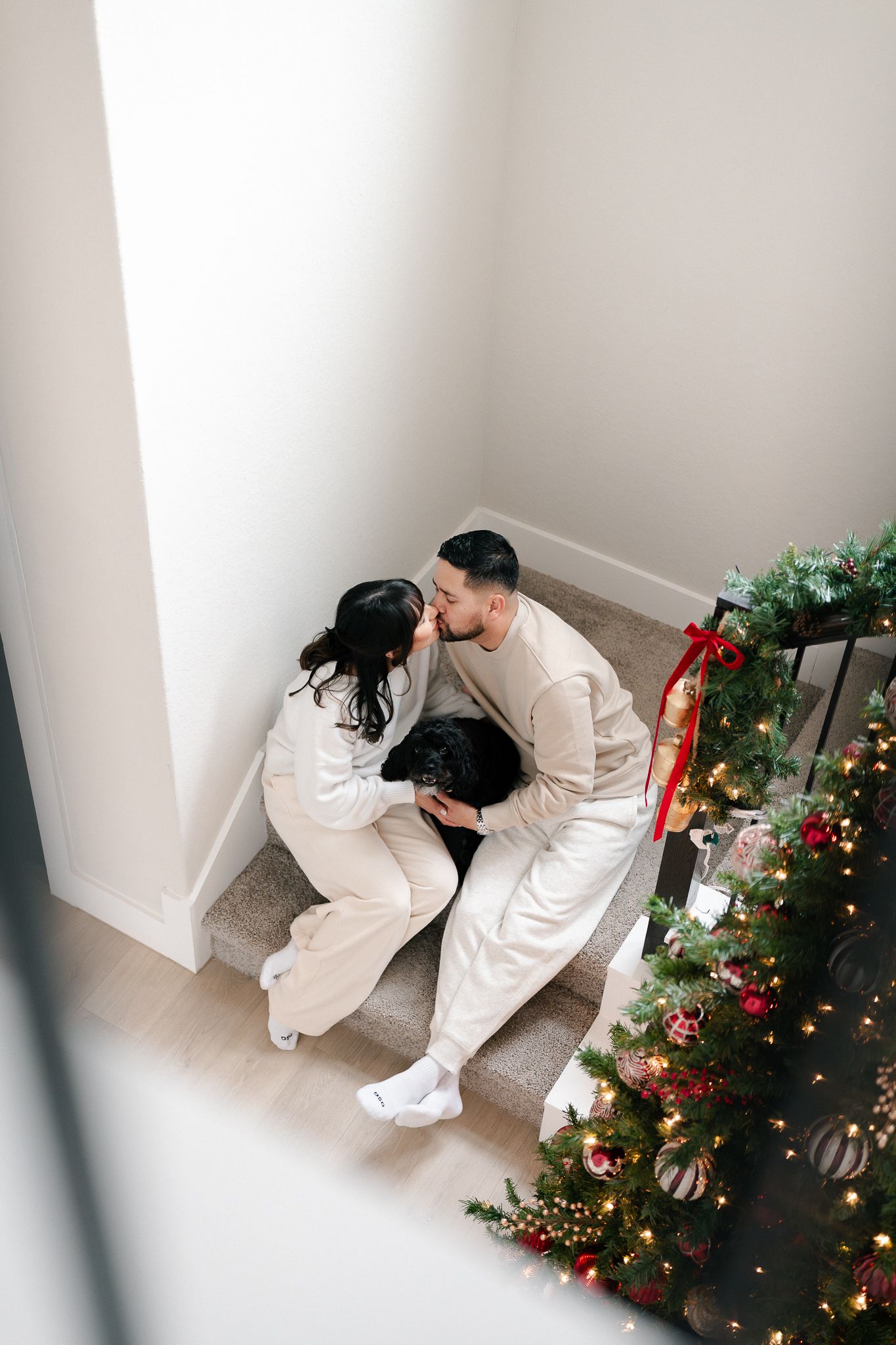 Couple in matching white outfits sitting on floor near decorated Christmas tree with lights and ornaments.