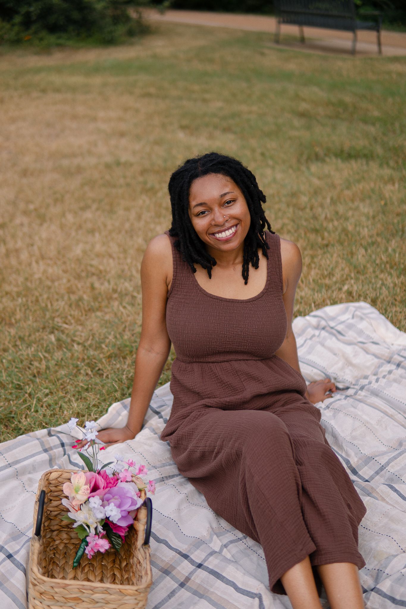 A picnic scene with a brown dress, blanket, and basket filled with colorful flowers on grass.