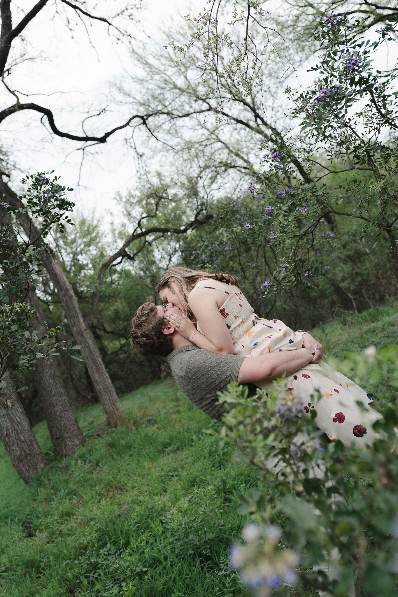 A couple sharing a romantic kiss in a lush green park surrounded by blooming trees and wildflowers.