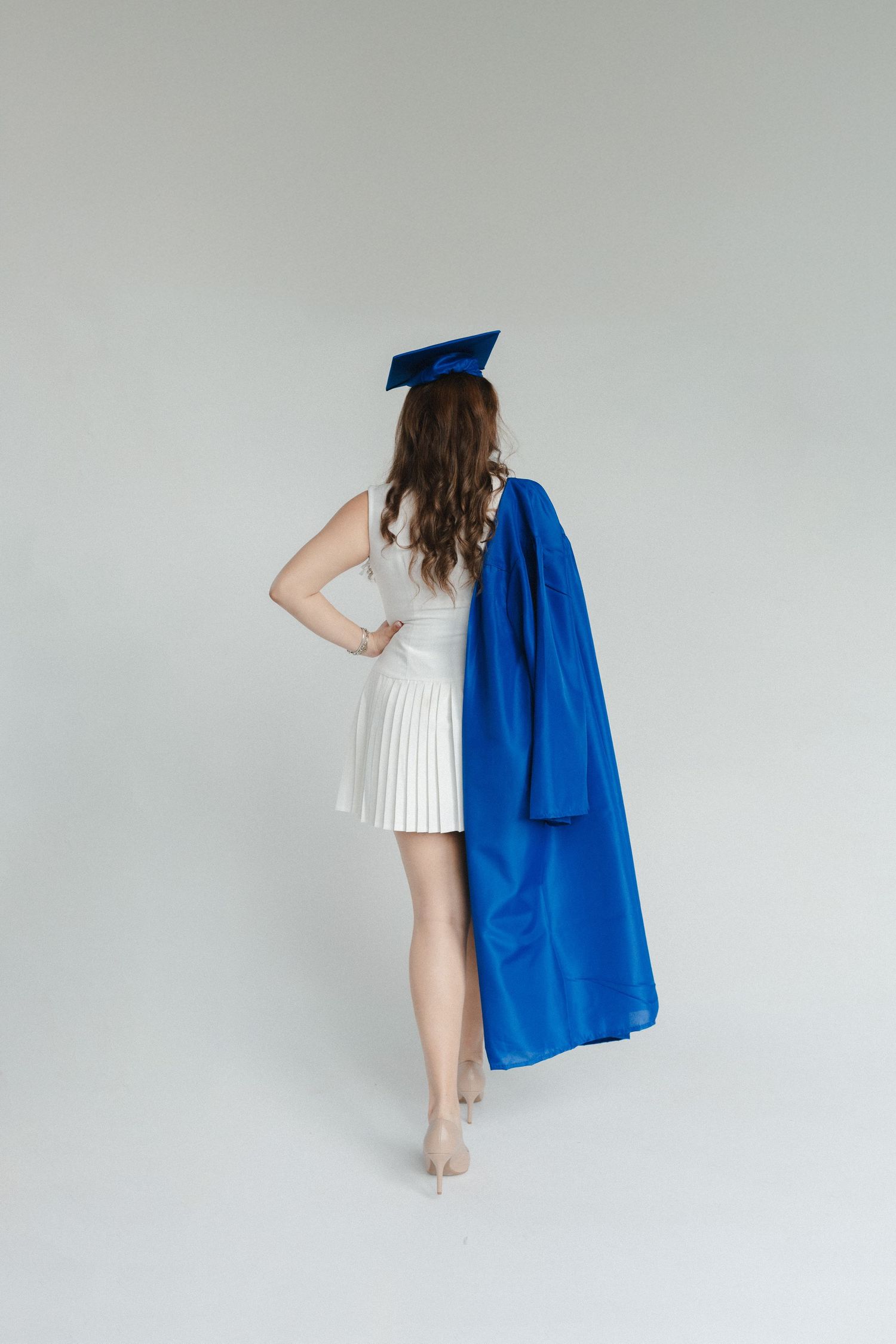 Young woman in white dress and blue graduation cap holding academic gown, viewed from behind on white background.