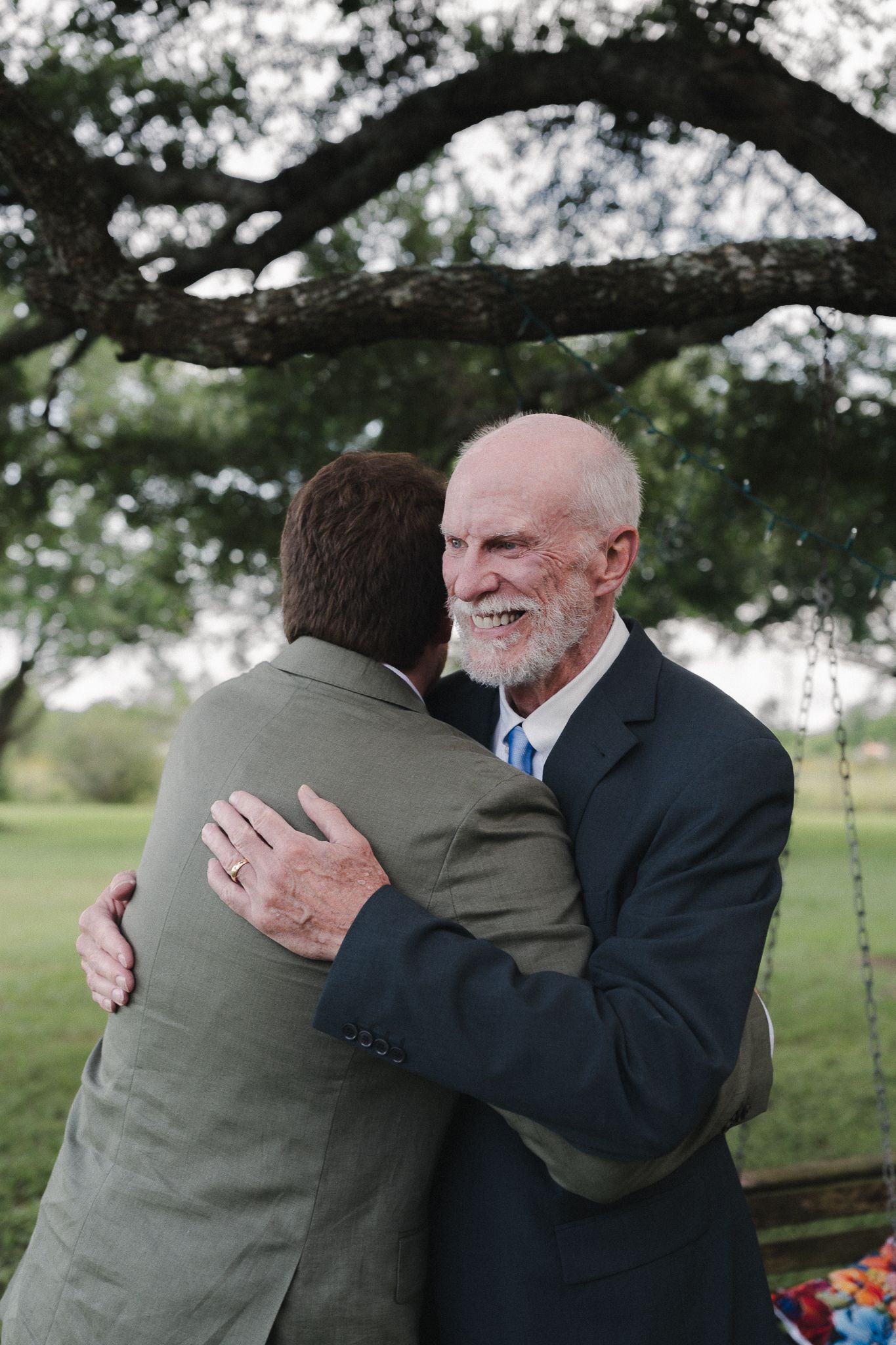 Two men share a warm embrace outdoors at a wedding, with an older smiling gentleman in a navy suit hugging a younger man.