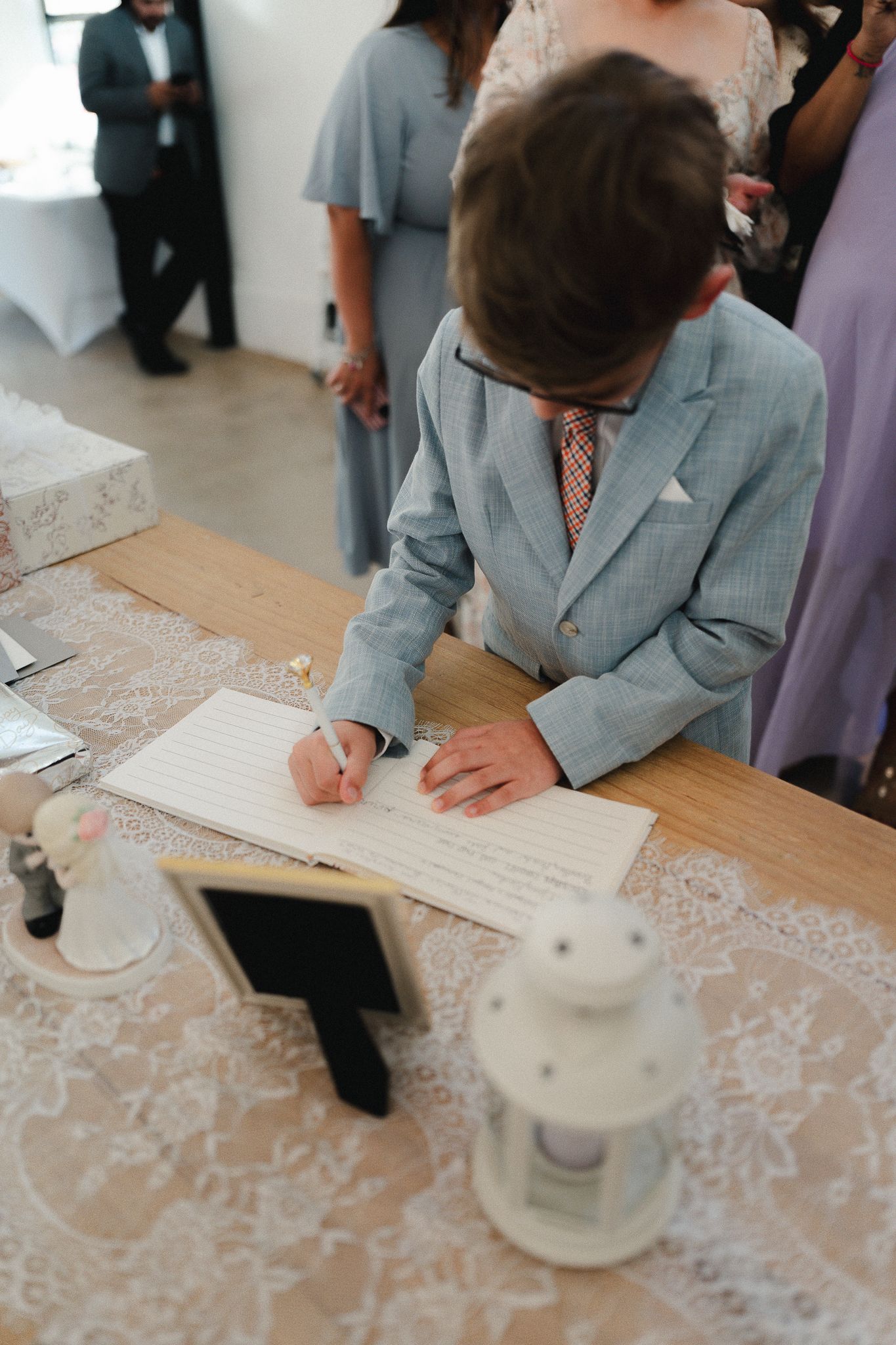 Young boy in gray suit signing wedding guest book at lace-covered table during wedding reception.