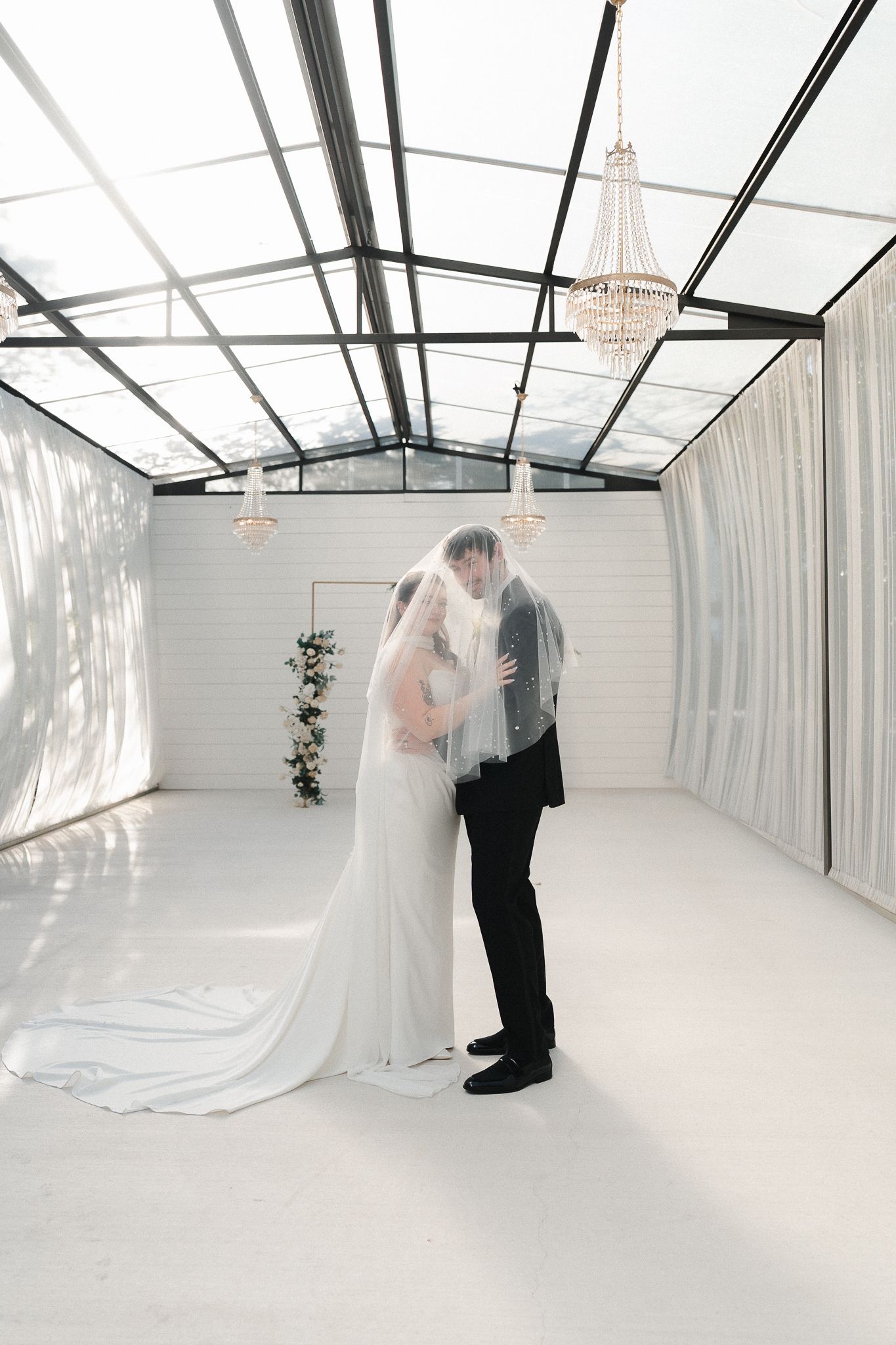 Bride and groom share a kiss under a flowing veil in an elegant white greenhouse wedding venue with crystal chandeliers.