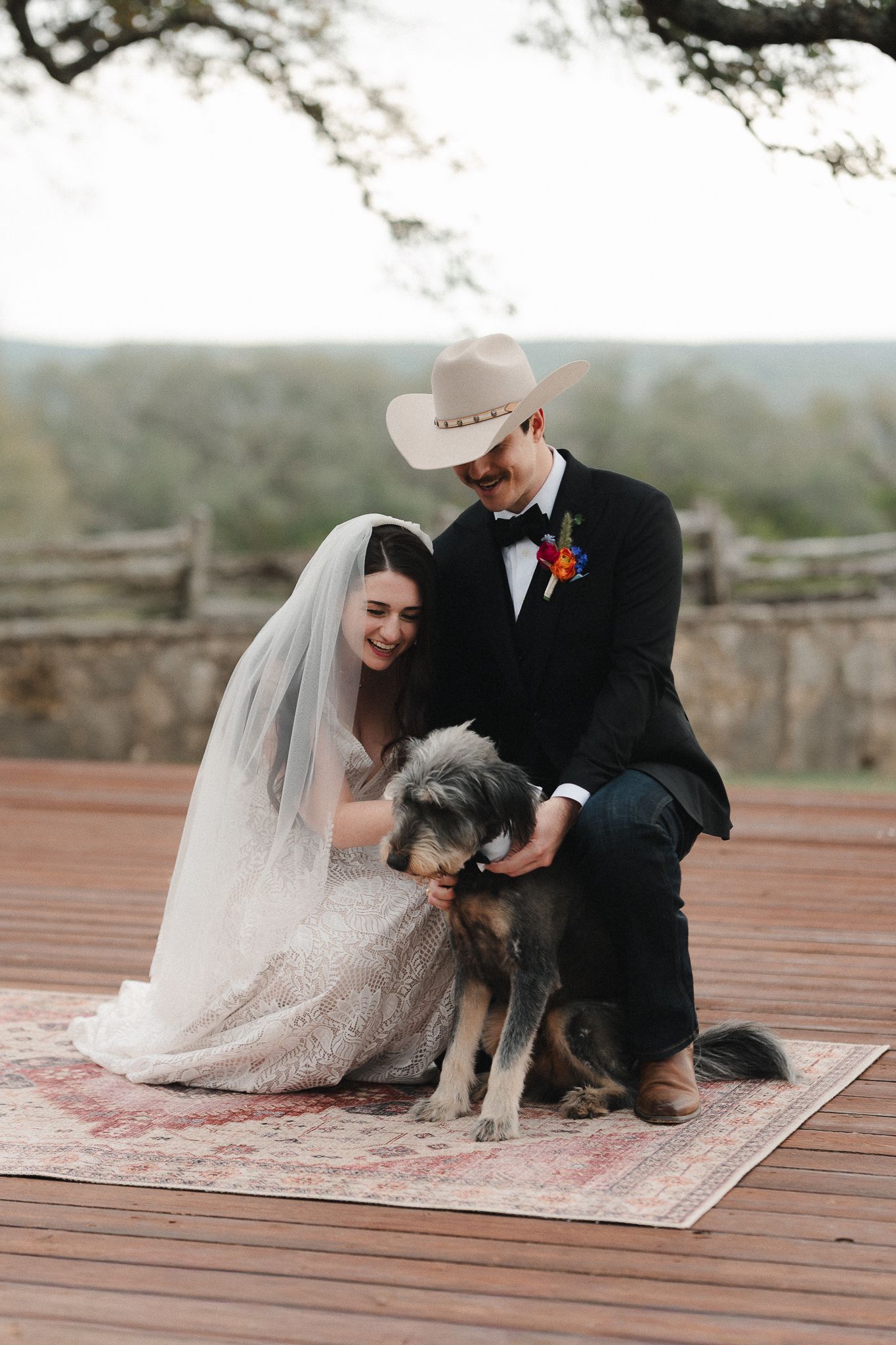Bride and groom in cowboy hat petting their dog on a wooden deck at an outdoor rustic wedding.